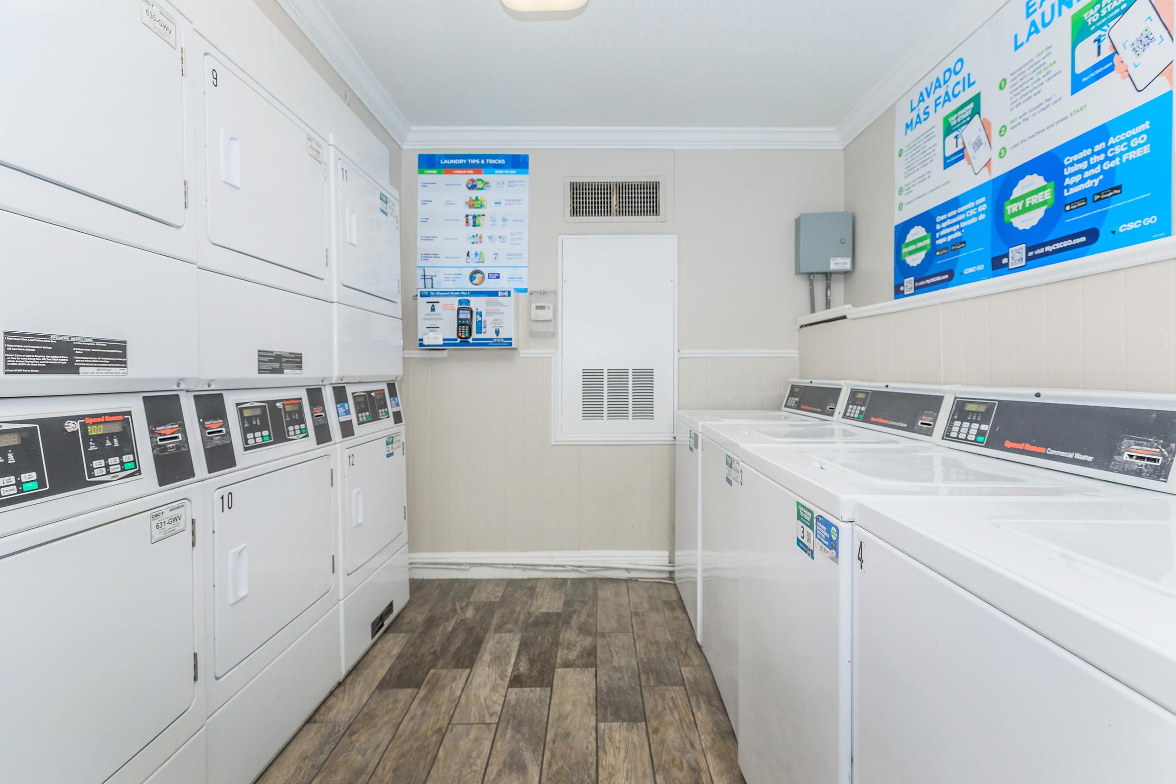 A clean laundry room featuring multiple white washing machines and dryers lined along the walls. There is a wall-mounted information board displaying laundry instructions and pricing. The room has a modern design with wood-style flooring and bright lighting.