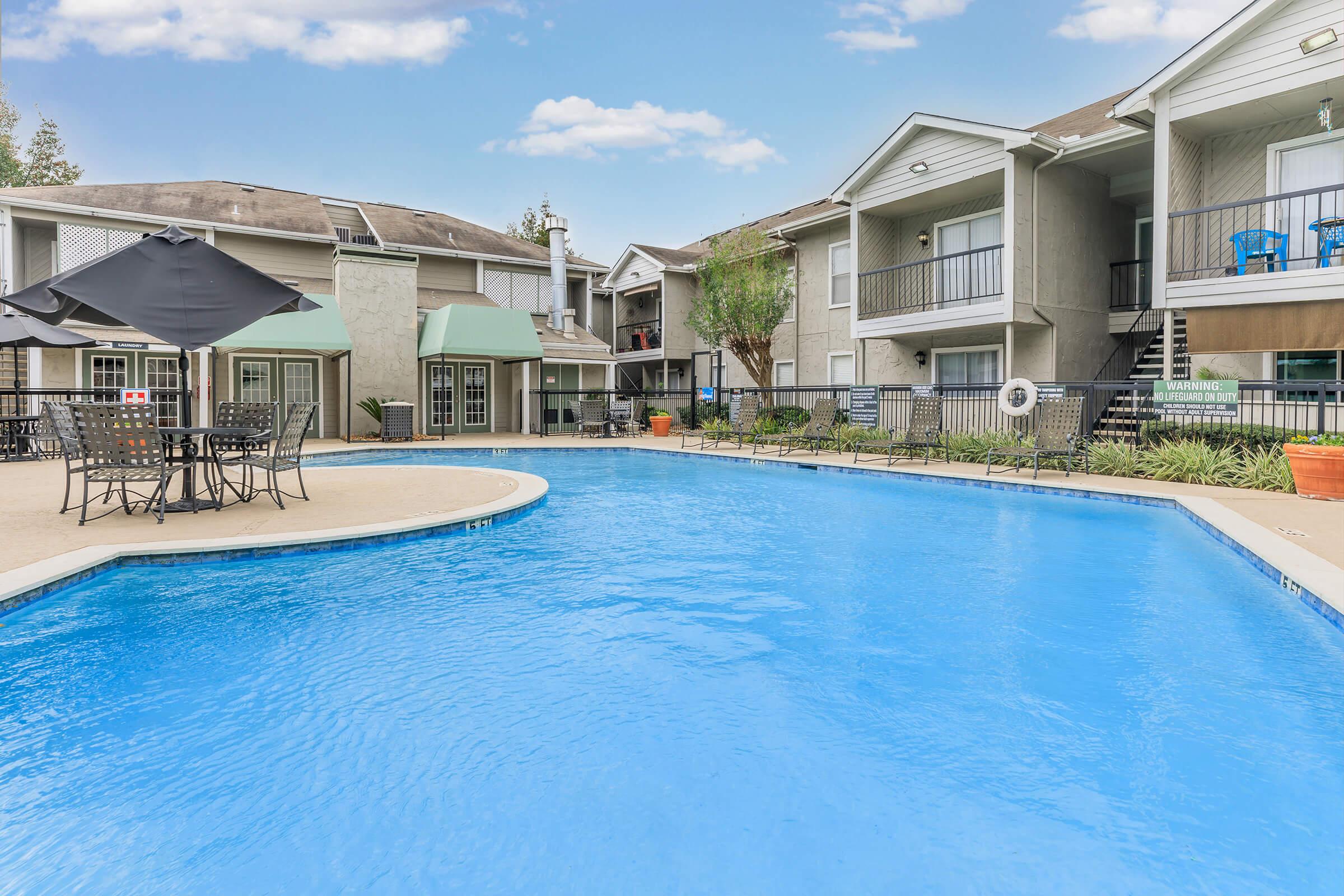 A bright blue swimming pool surrounded by lounge chairs and a table with an umbrella, set in a landscaped courtyard featuring apartment buildings in the background. The scene is inviting and calm, perfect for relaxation and leisure. Clear skies reflect above the pool.
