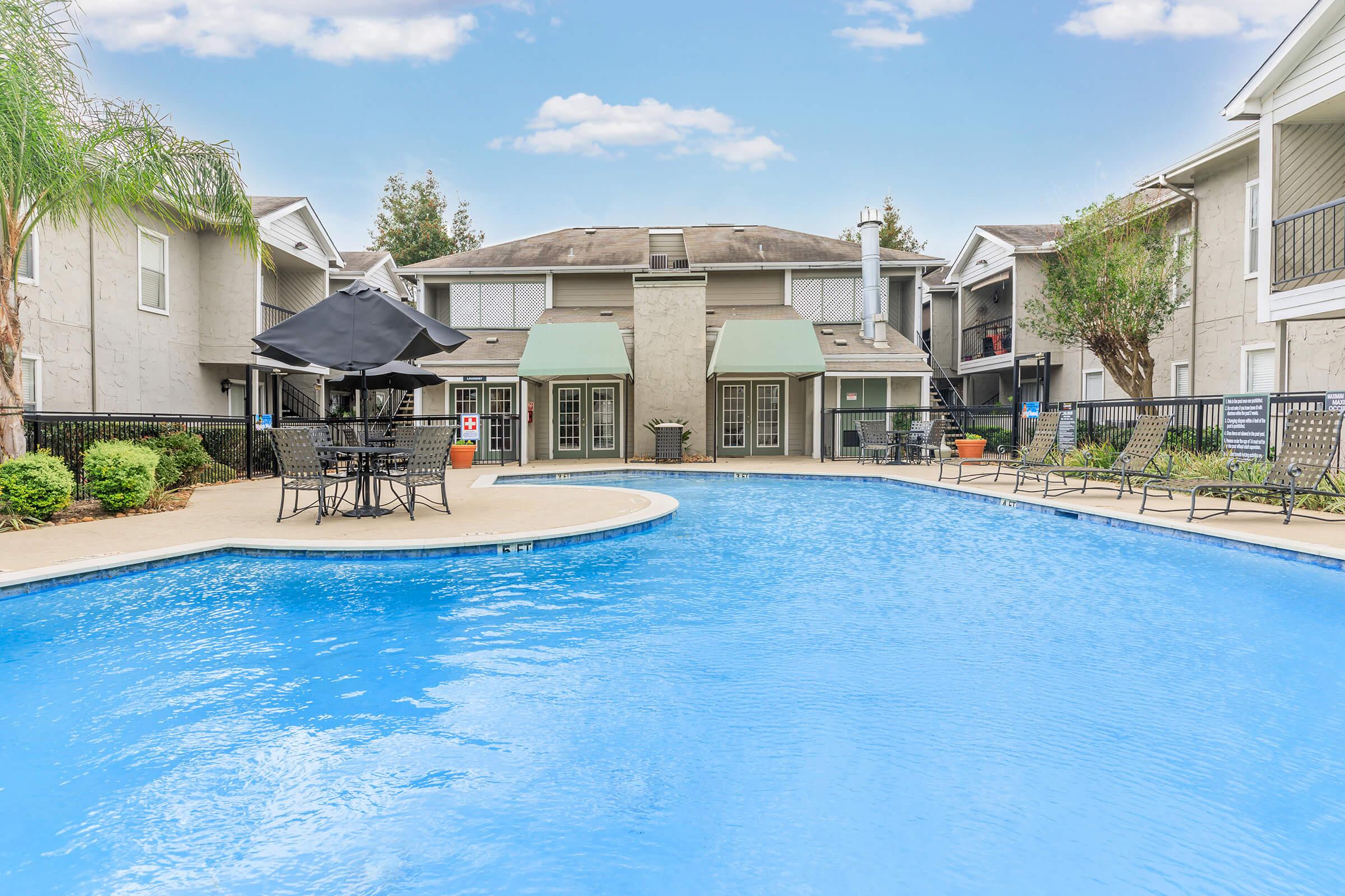 A bright, inviting swimming pool surrounded by lounge chairs and patio tables. In the background, a residential building features awnings and greenery, set against a clear blue sky. The pool area is well-maintained, offering a relaxing atmosphere.