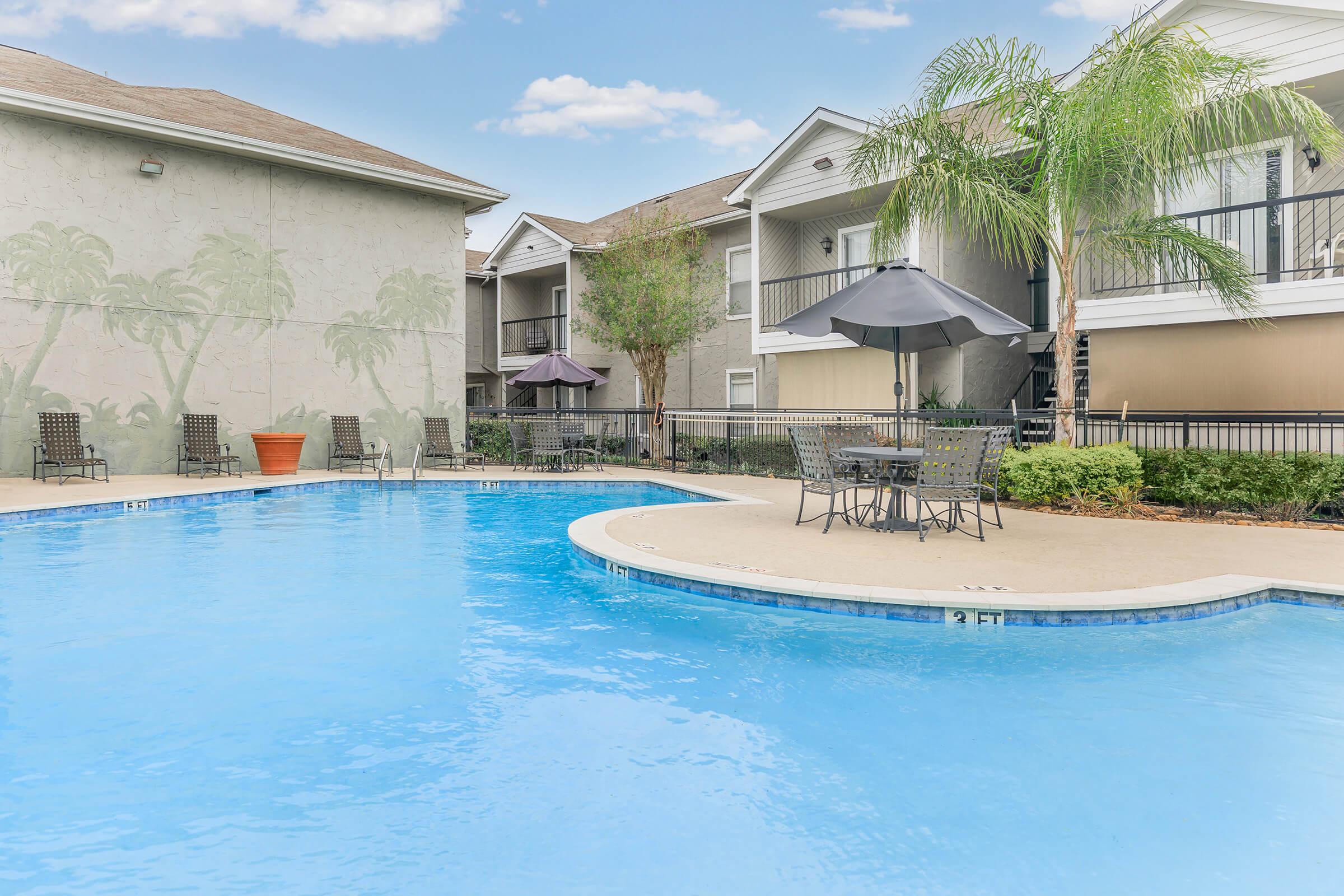 A clear blue pool surrounded by lounge chairs and umbrellas, with apartment buildings in the background. Palm trees are depicted on the wall, adding a tropical feel to the scene. The area is well-maintained, creating a relaxing atmosphere for residents and guests.