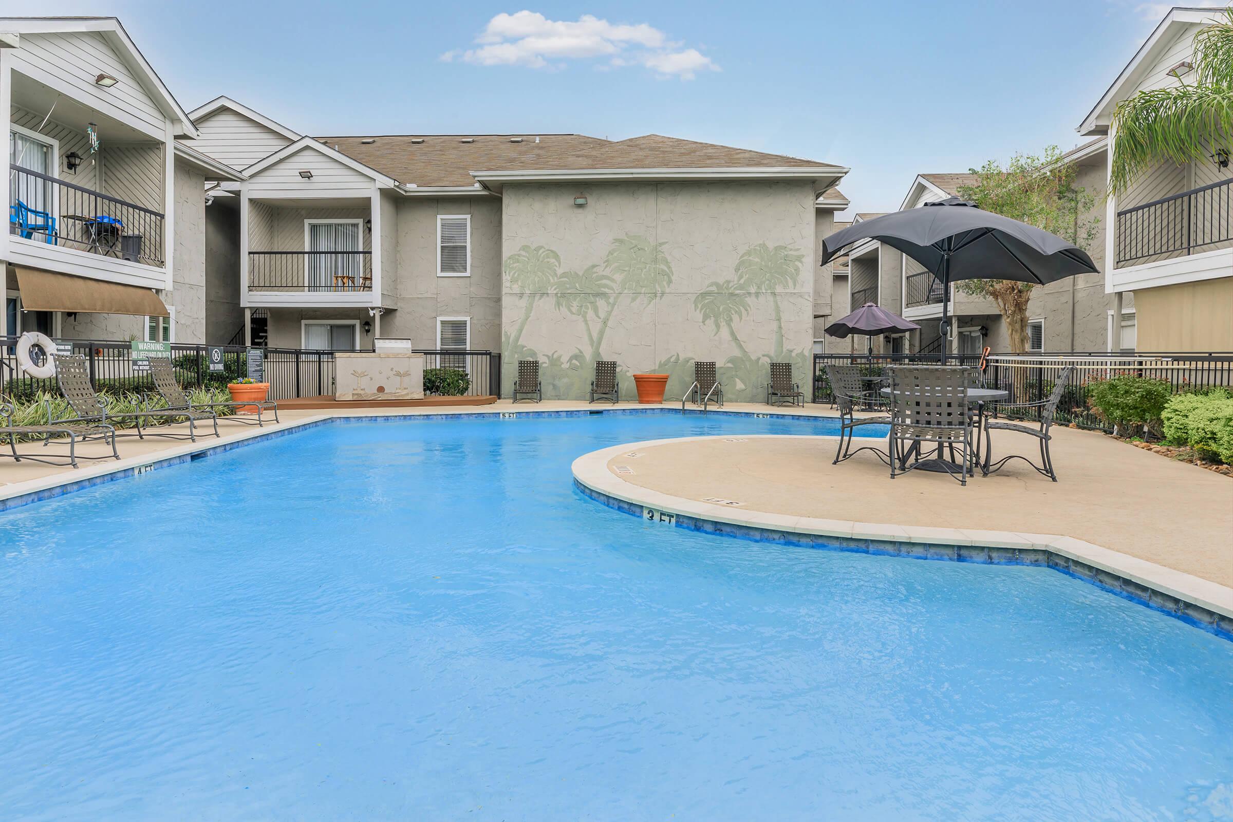 A spacious outdoor swimming pool surrounded by lounge chairs and tables. Umbrellas provide shade in a relaxing pool area, with a building and palm tree mural in the background. The setting is inviting and perfect for leisure and relaxation. Clear blue sky above enhances the serene atmosphere.