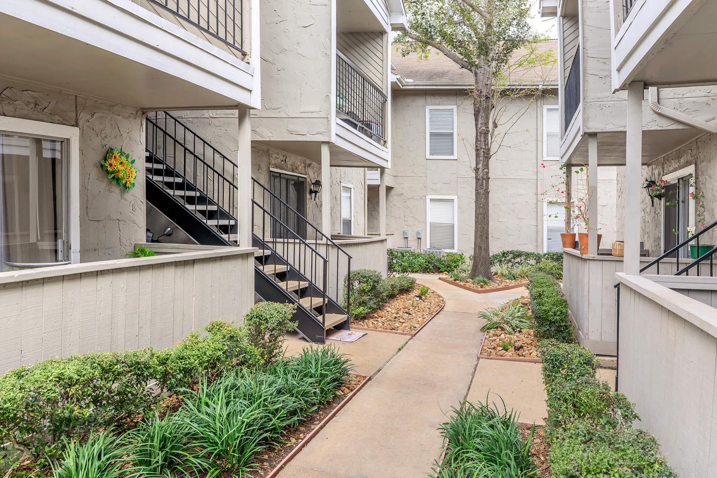 A peaceful courtyard view of an apartment complex featuring a walking path lined with manicured shrubs and small rocks. On the left, a staircase leads to upper-level apartments, while the surrounding buildings are painted in light gray. Greenery adds a touch of color to the serene environment.