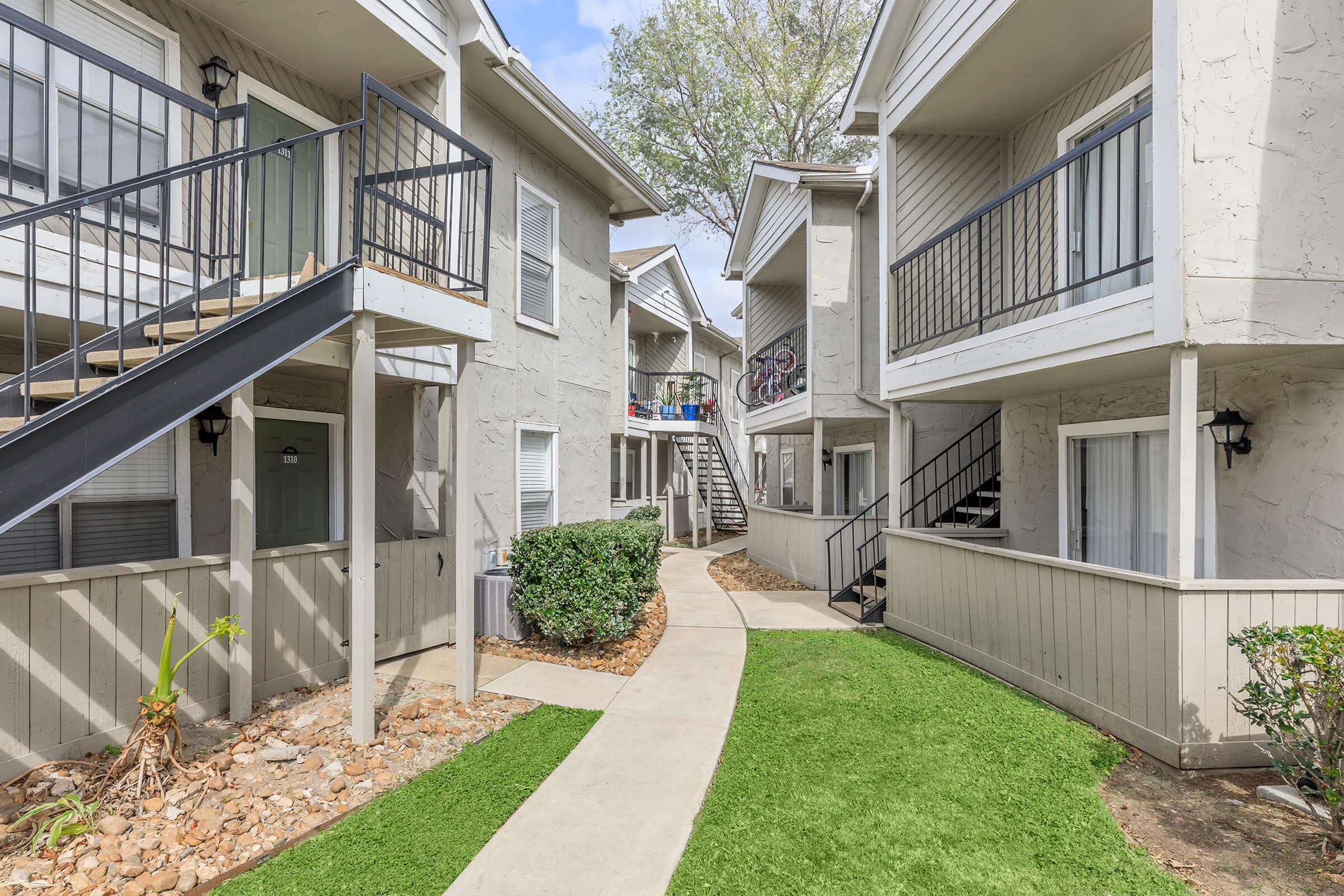 A view of a multi-unit apartment complex with beige stucco walls. Concrete walkways and grassy areas lead between the buildings, which have balconies and staircases. The environment appears well-maintained, with landscaping elements like small bushes and stones.