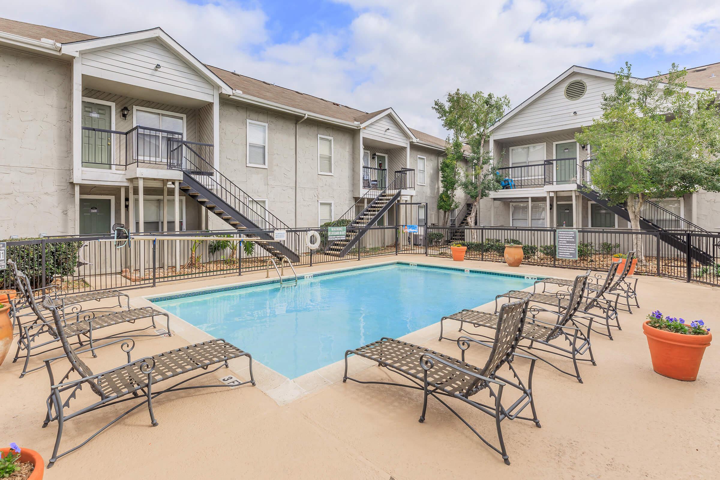 A tranquil apartment complex courtyard featuring a rectangular swimming pool surrounded by several lounge chairs. Greenery and potted plants add to the inviting atmosphere. Two-story buildings flank the pool area, with balconies visible on the upper level. Cloudy sky overhead.