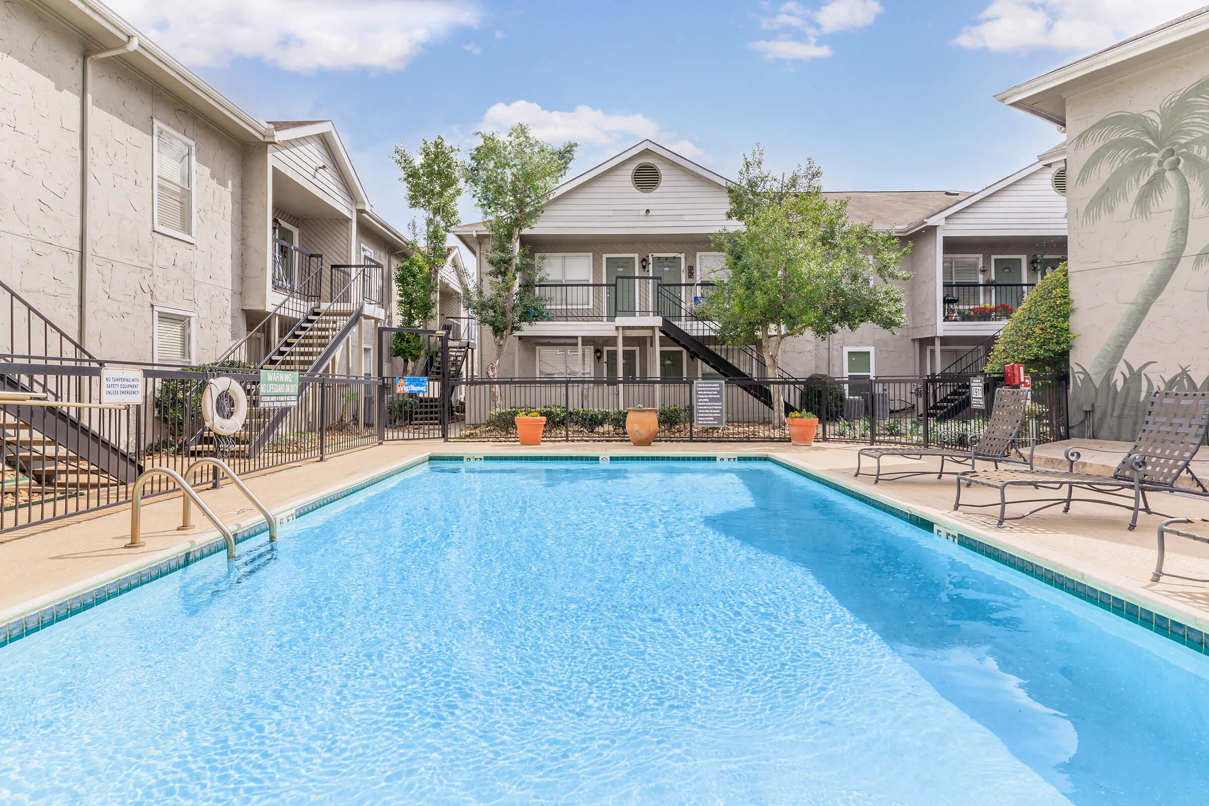 A sparkling blue swimming pool surrounded by lounge chairs and potted plants, with residential buildings in the background. The area features stairs leading to upper-level apartments, and palm trees add a tropical touch to the serene atmosphere. Bright sky with fluffy clouds completes the scene.