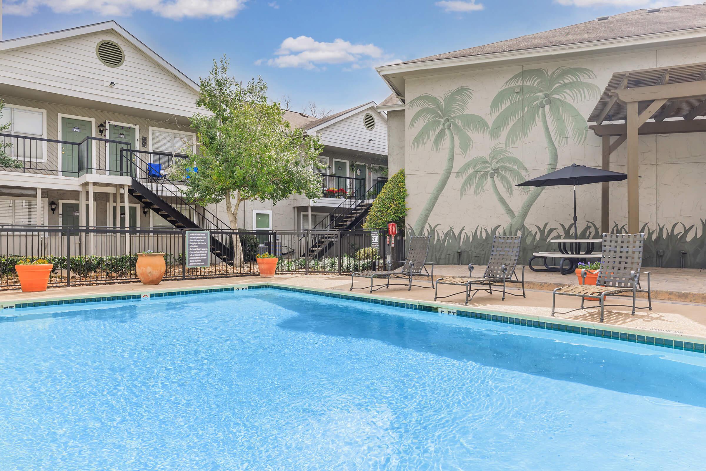 A sunny pool area featuring a clear blue swimming pool surrounded by lounge chairs and potted plants. In the background, there are two-story apartment buildings with a mural of palm trees. A shaded seating area with a picnic table and umbrella is also visible. The overall setting conveys a relaxing outdoor atmosphere.