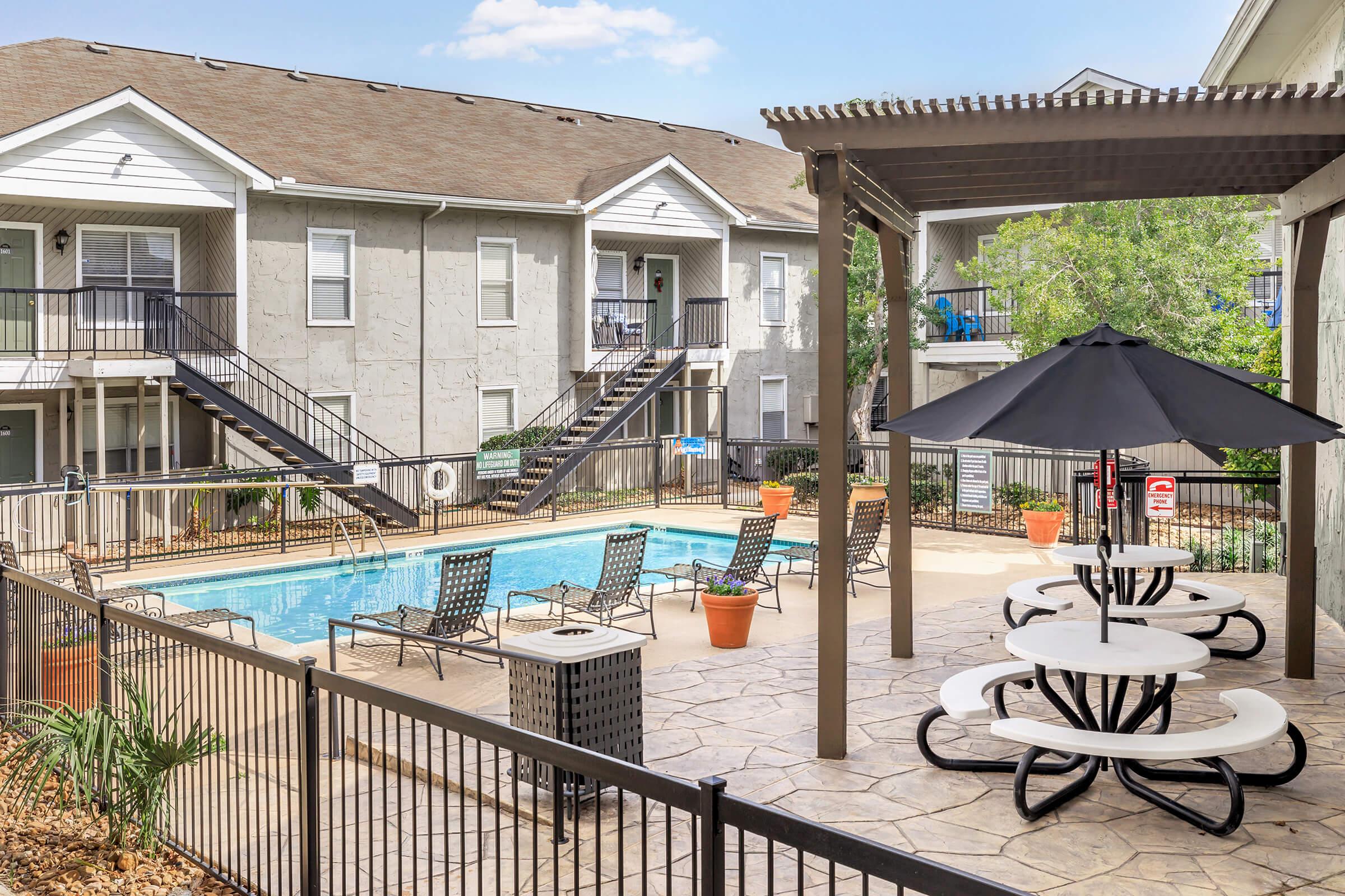 A community pool area featuring a clear blue pool surrounded by lounge chairs and tables with umbrellas. The area is enclosed by a fence and bordered by well-kept landscaping. In the background, there are two apartment buildings with staircases leading to the upper levels.