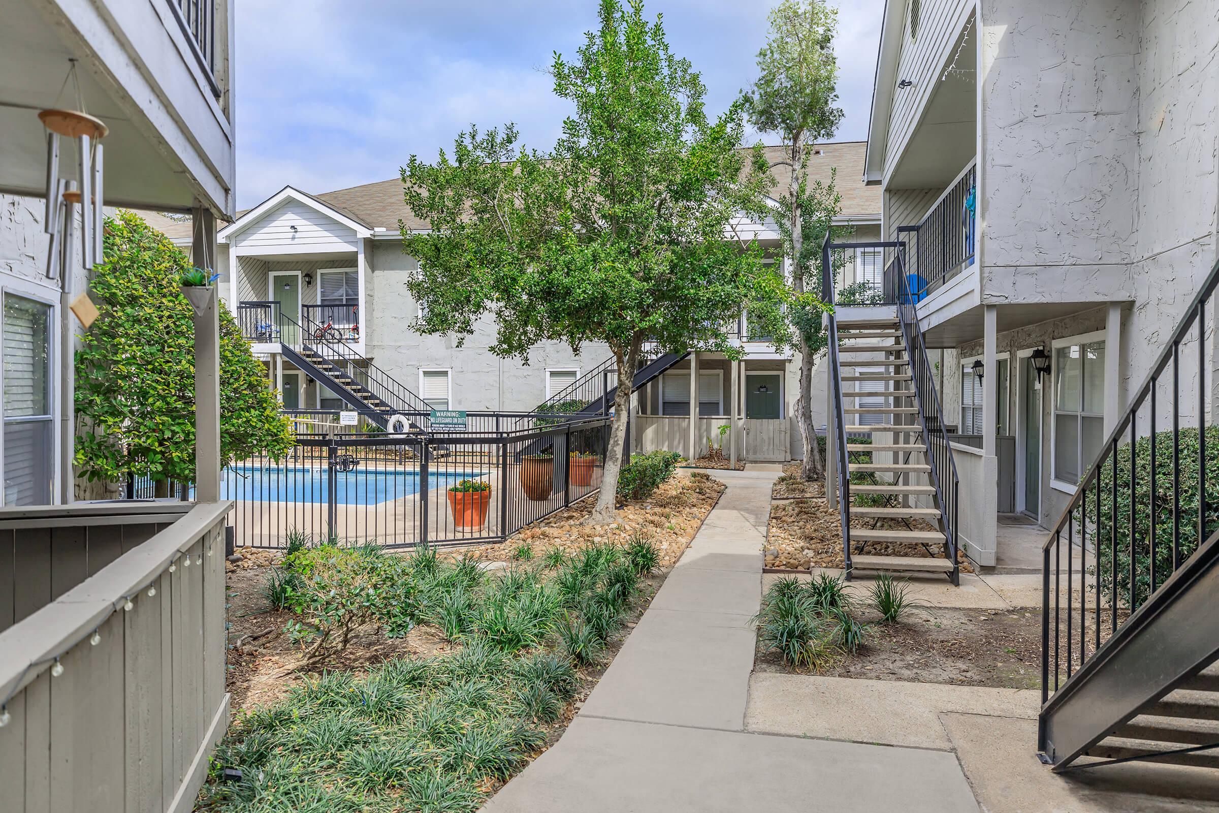 A view of a peaceful courtyard between two apartment buildings, featuring a pathway lined with greenery and a tree. In the background, there's a swimming pool surrounded by a wooden fence. The sky is partly cloudy, creating a serene atmosphere perfect for relaxation.