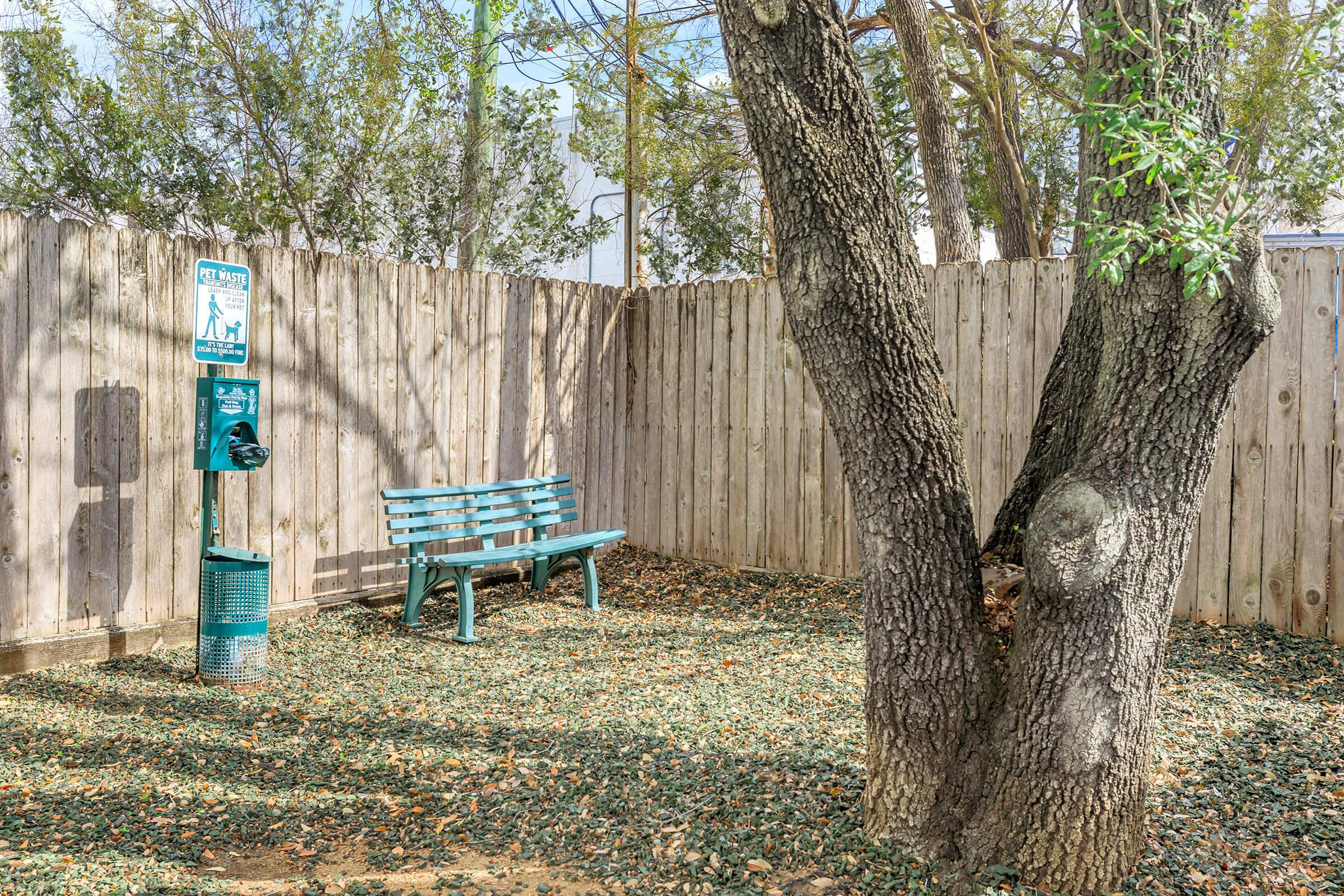 A quiet corner of a park featuring a green bench beside a large tree, surrounded by a bed of leaves. There is a waste disposal station for pet waste nearby, set against a wooden fence and a clear sky.
