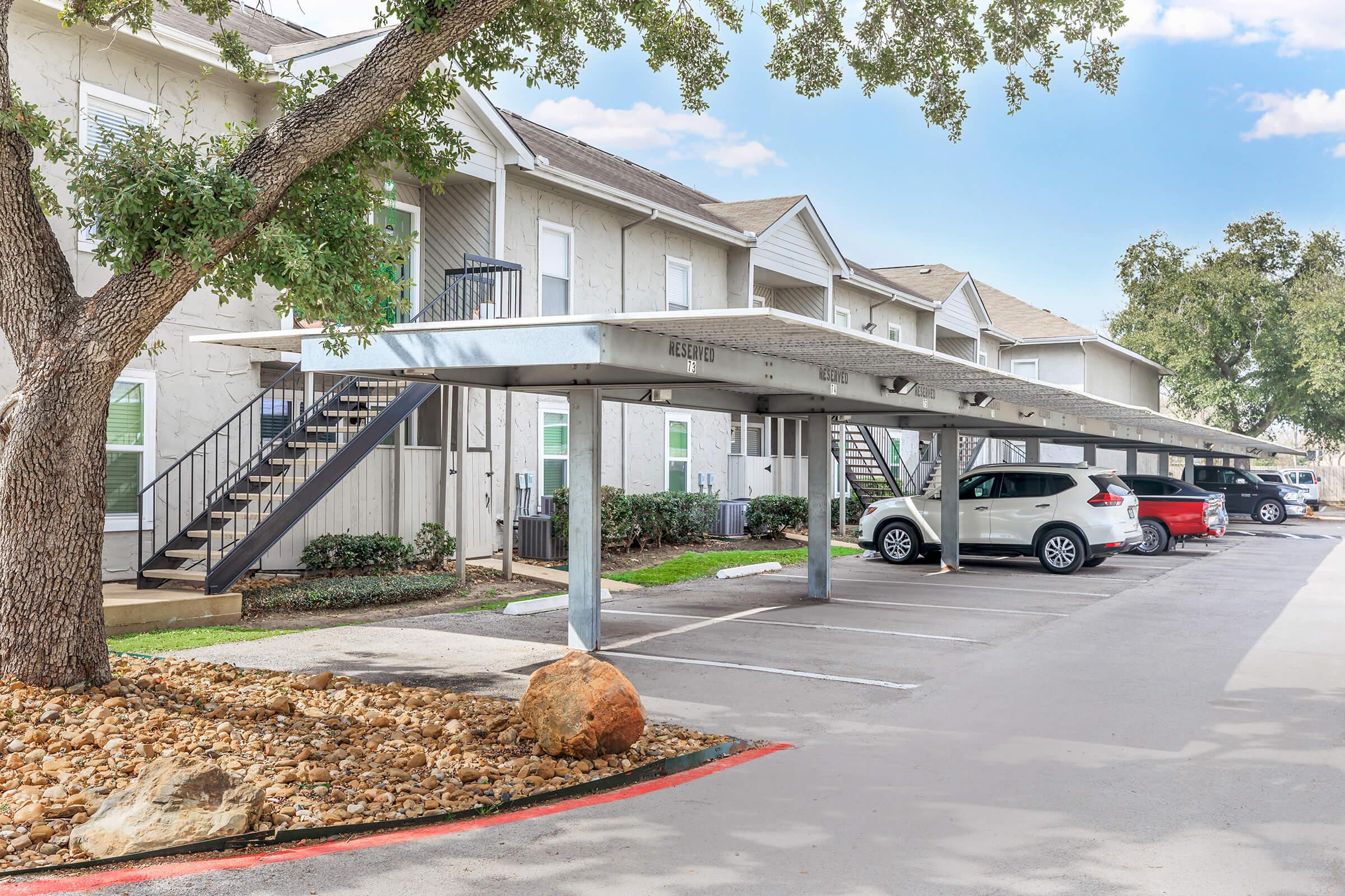 A parking area with covered spaces and rocks, adjacent to a two-story apartment building. The building features exterior stairs leading to upper units, surrounded by trees and a clear blue sky. Several vehicles are parked under the cover.