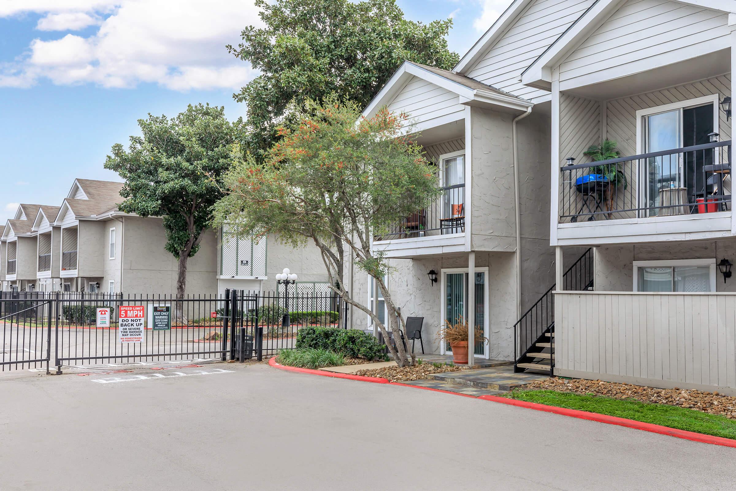 Exterior view of a modern residential complex featuring two-story buildings with balconies. A well-maintained walkway leads to a gated entrance, surrounded by lush greenery and trees. Parking spaces are visible in front of the gated area, and a clear blue sky adds to the pleasant atmosphere.