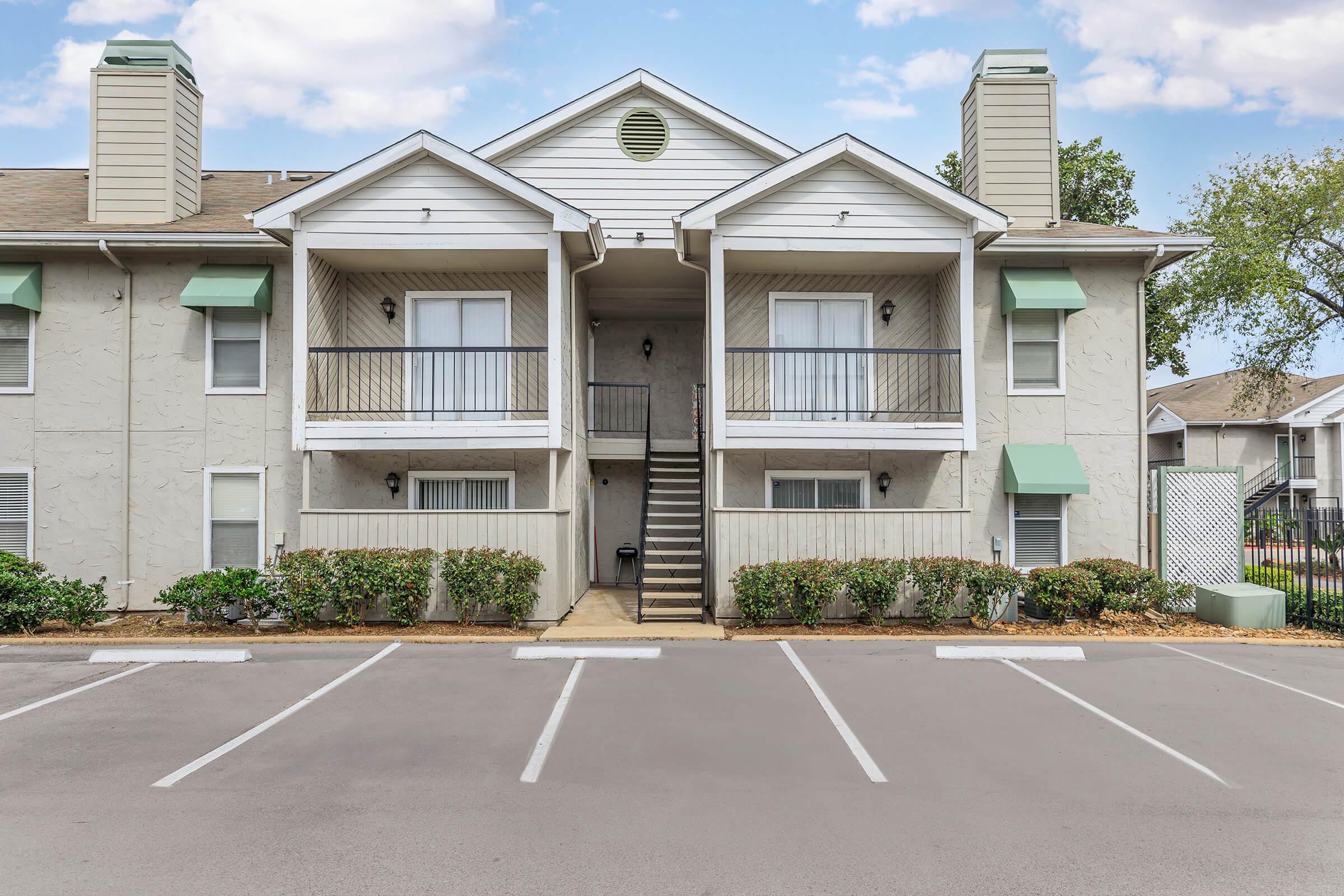 A two-story apartment building with a light gray exterior. The building has two front balconies with white railings, flanked by green awnings. A central staircase leads to the upper units. The parking area in front is lined with painted white lines, and well-maintained shrubs surround the building.