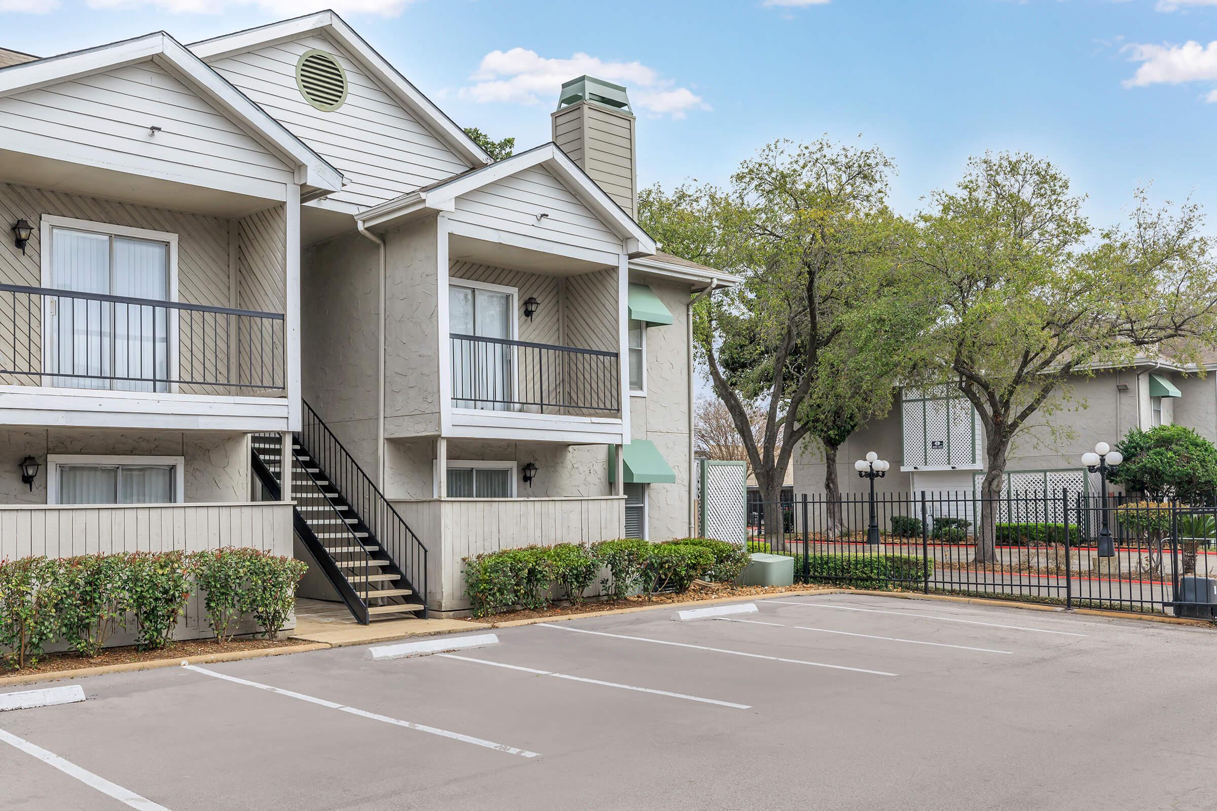 Exterior view of a two-story residential building with a staircase leading to a balcony. The image features landscaped greenery, including trees and hedges, alongside a parking lot with empty spaces. A gated area is visible in the background with additional buildings.