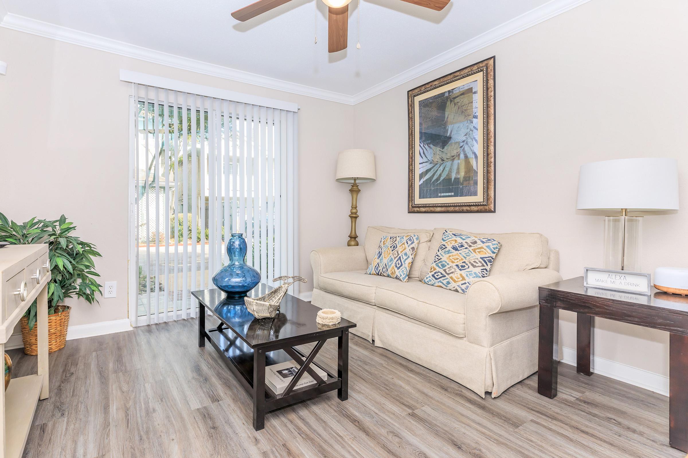 A cozy living room featuring a light beige couch with patterned pillows, a dark wooden coffee table, a decorative blue vase, and a floor lamp. Sliding glass doors with blinds provide natural light, while a green plant adds a touch of nature to the space.