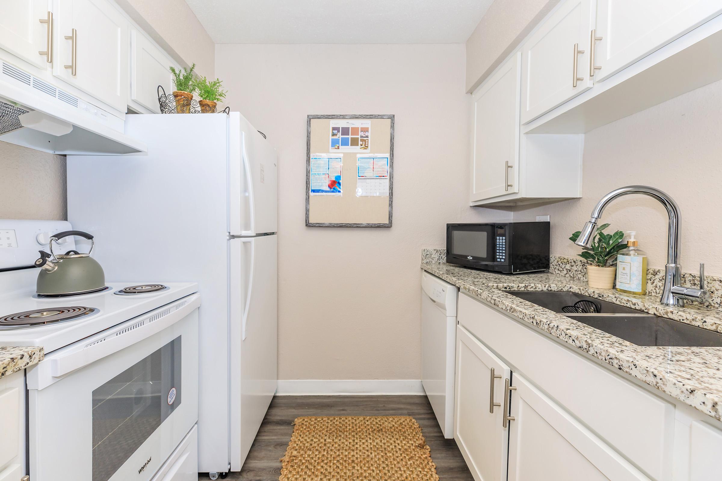 A modern kitchen featuring white cabinetry, a granite countertop, a stove, oven, and refrigerator. A microwave is on the counter, alongside a sink with a faucet. The wall displays a framed bulletin board. A woven mat is laid on the floor, adding a cozy touch to the space.