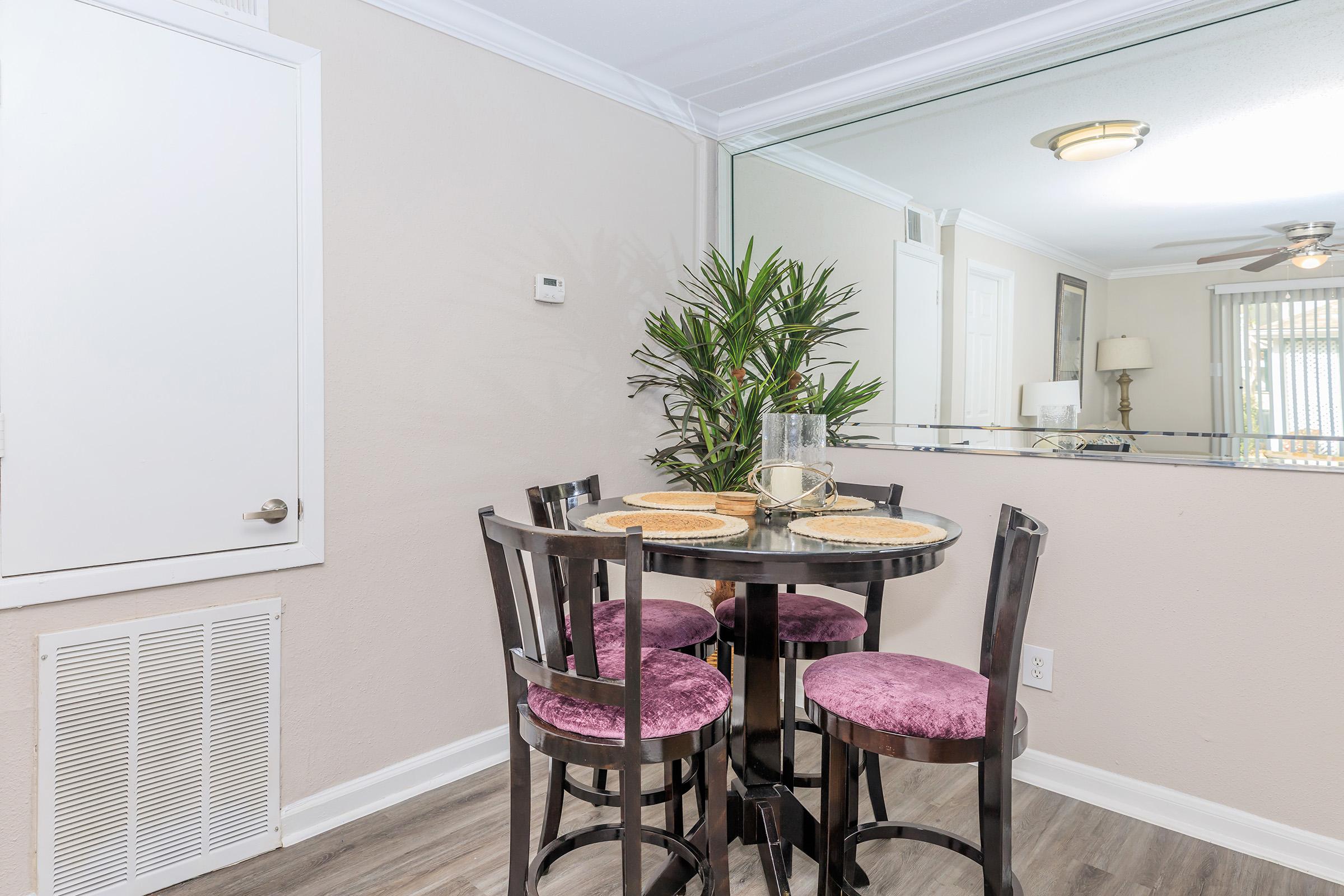 A small dining area featuring a round table with four purple upholstered chairs. The table is set with beige placemats and a decorative glass vase. A mirror reflects the light in the room, and a potted plant adds a touch of greenery. Light-colored walls and laminate flooring create a modern, cozy atmosphere.