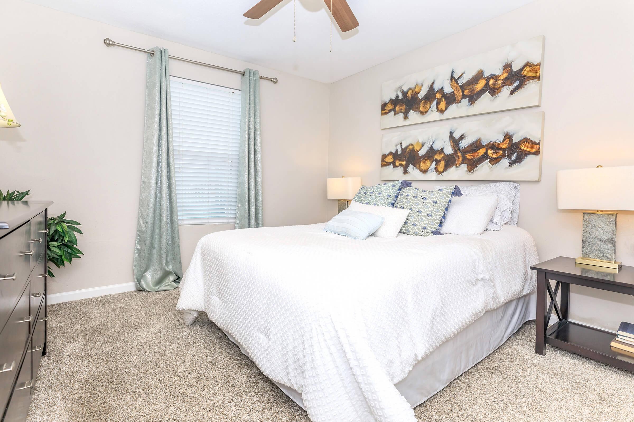 A cozy bedroom with a neatly made bed featuring a white comforter and various decorative pillows. There are two lamps on nightstands beside the bed, and a large piece of abstract wall art above the headboard. Light curtains frame a window, and a potted plant adds a touch of greenery to the space.