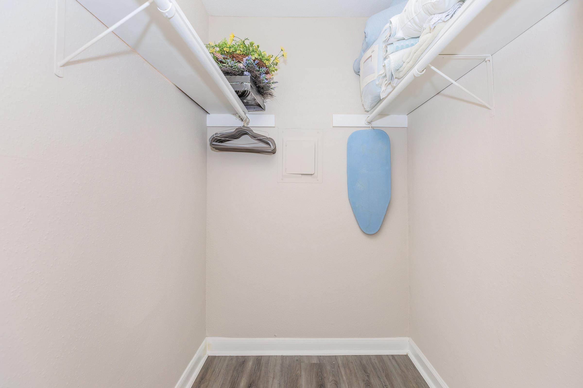 A small, organized closet featuring two hanging shelves. The upper shelf holds a potted plant and a neatly folded blanket, while the lower section has a few hangers and a blue ironing board attached to the wall. The walls are painted light, and the flooring is a wood-like laminate.