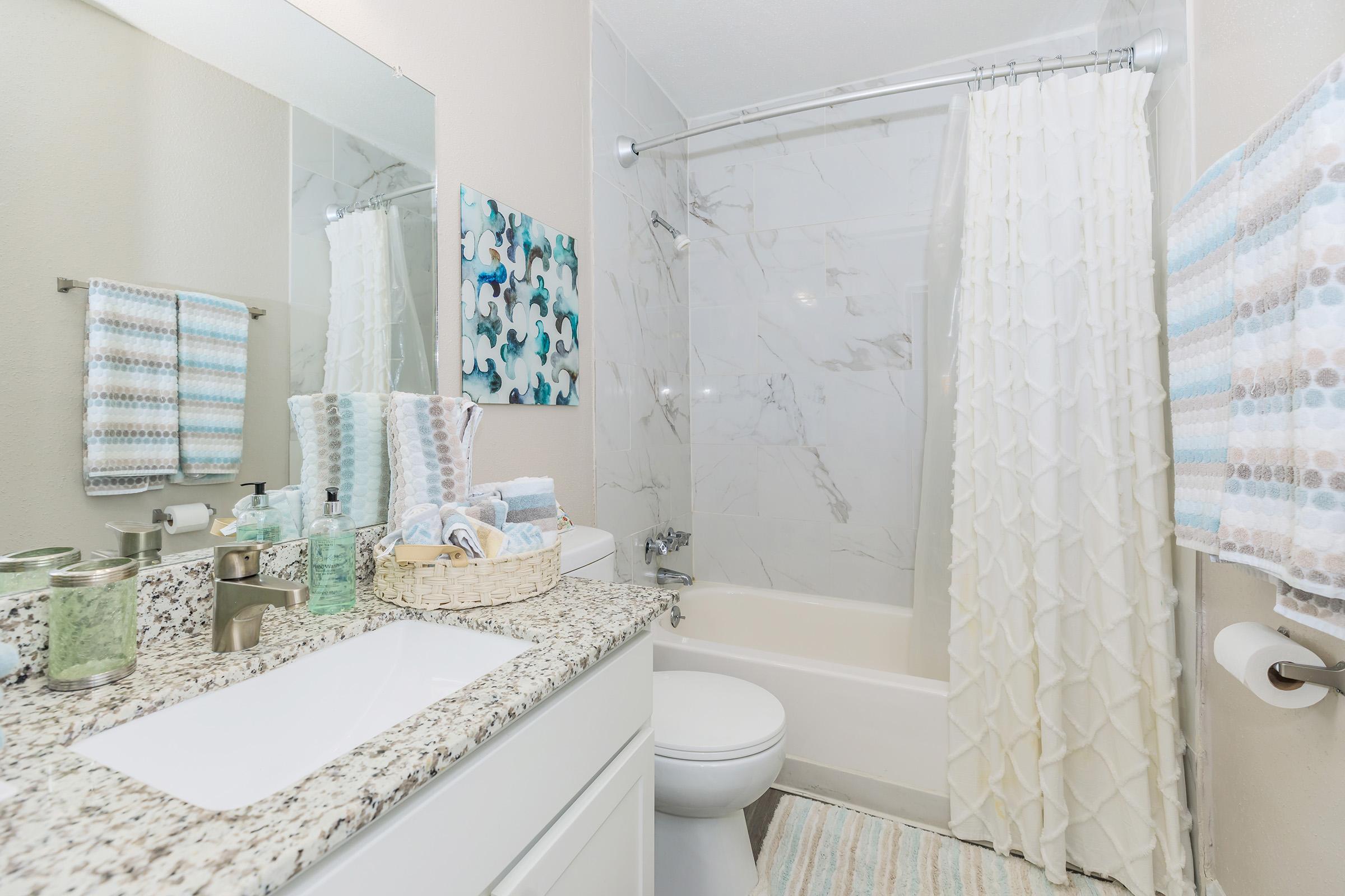 A modern bathroom featuring a white bathtub and shower with a marble wall, a granite countertop with a sink, and decorative towels. The space is well-lit with a large mirror reflecting the decor, including a colorful wall art piece. Soft pastel colors dominate the design, creating a serene atmosphere.