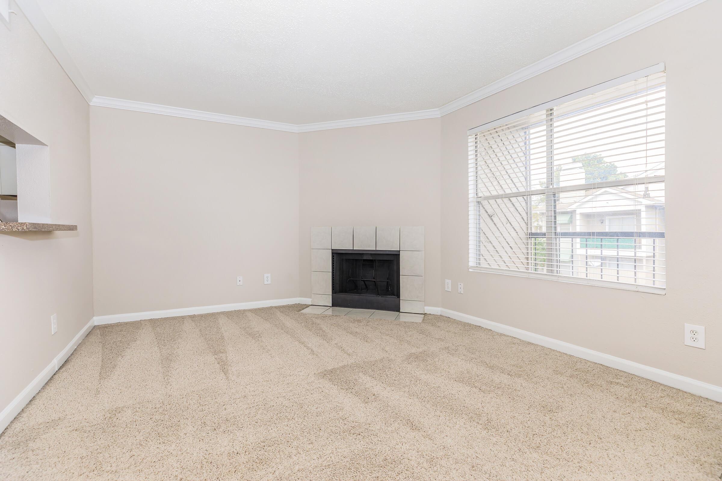 A spacious, empty living room featuring a light beige carpet, neutral-colored walls, and a fireplace with a tiled surround. The room has large windows with blinds, allowing natural light to enter. There is also a small opening leading to another area, suggesting an open layout.