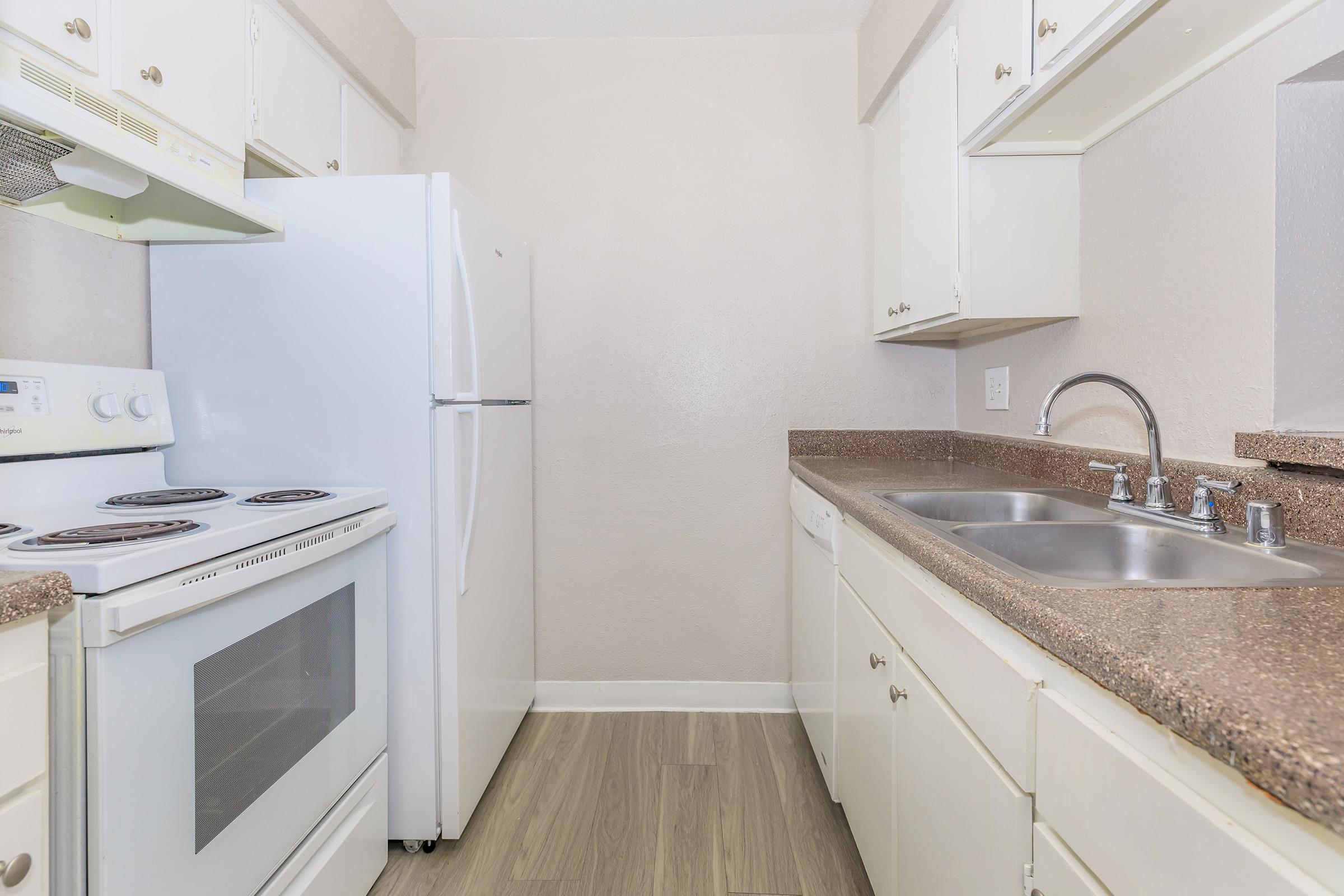 A small, modern kitchen featuring white cabinets, a white refrigerator, an electric stove with oven, and a double sink. The countertops are a light brown color, and the flooring is a light wood-like material. Walls are painted in a soft beige. The space is well-lit and clean.
