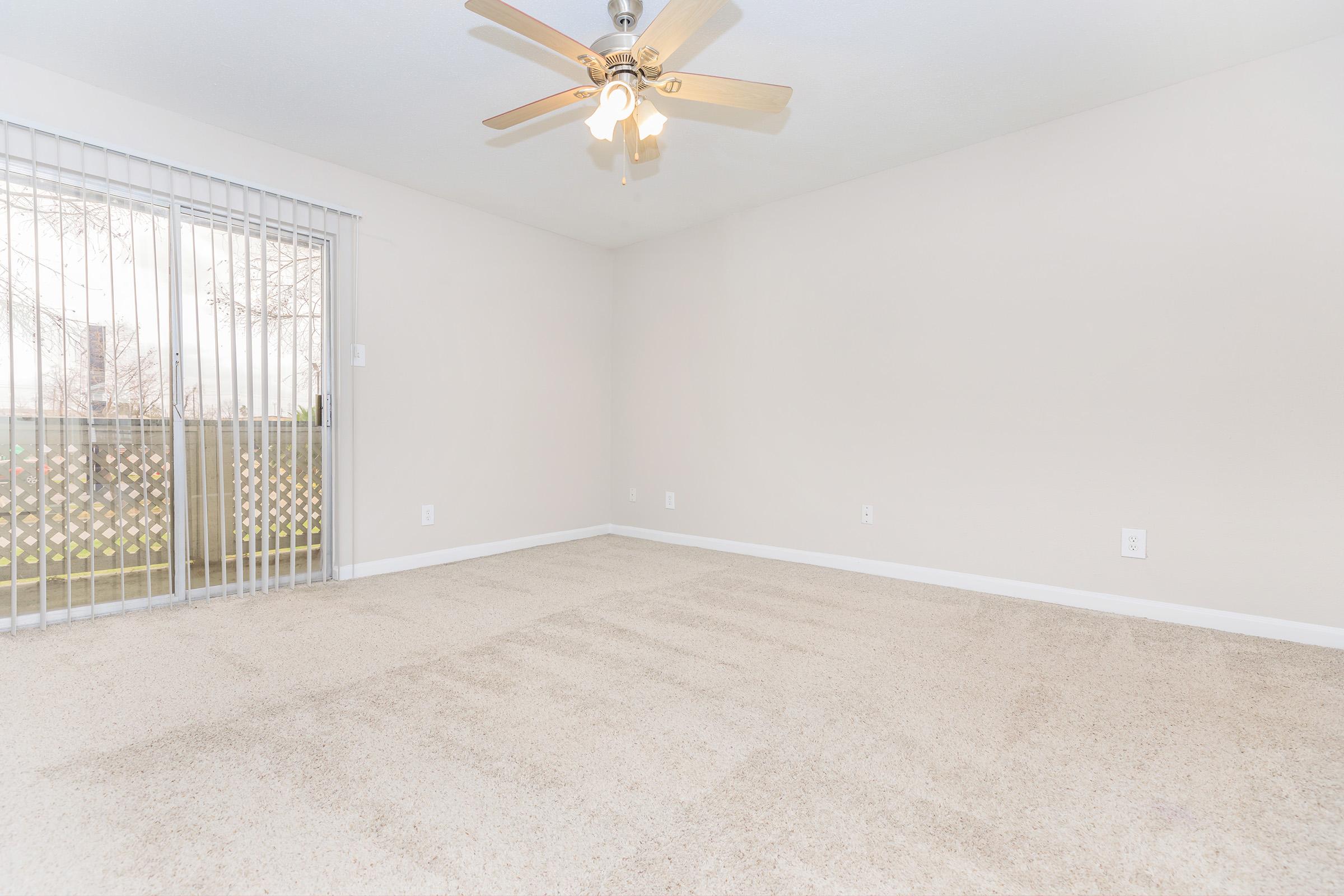 A spacious, empty room with light beige walls and a ceiling fan. The floor is covered in light carpet, and there is a sliding glass door leading to an outdoor area, surrounded by a wooden railing. Natural light illuminates the room, giving it a bright and airy feel.