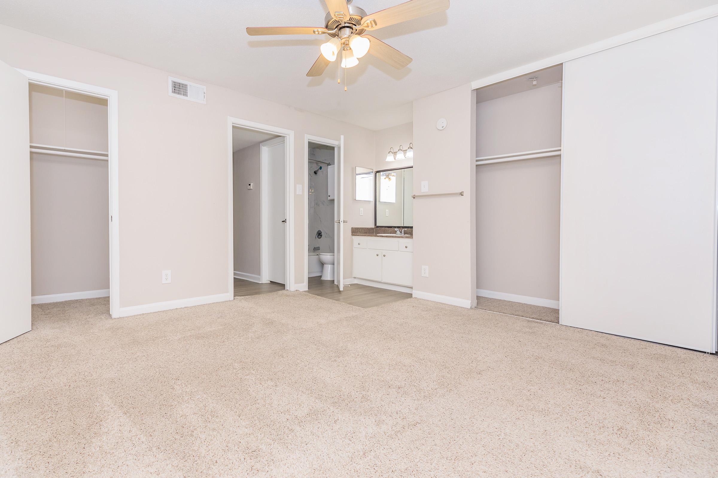 A spacious, empty room with light beige carpet, a ceiling fan, and a neutral color scheme. There are two closed closet doors on either side, a bathroom doorway visible in the back, and a small vanity area with a mirror integrated into the wall.