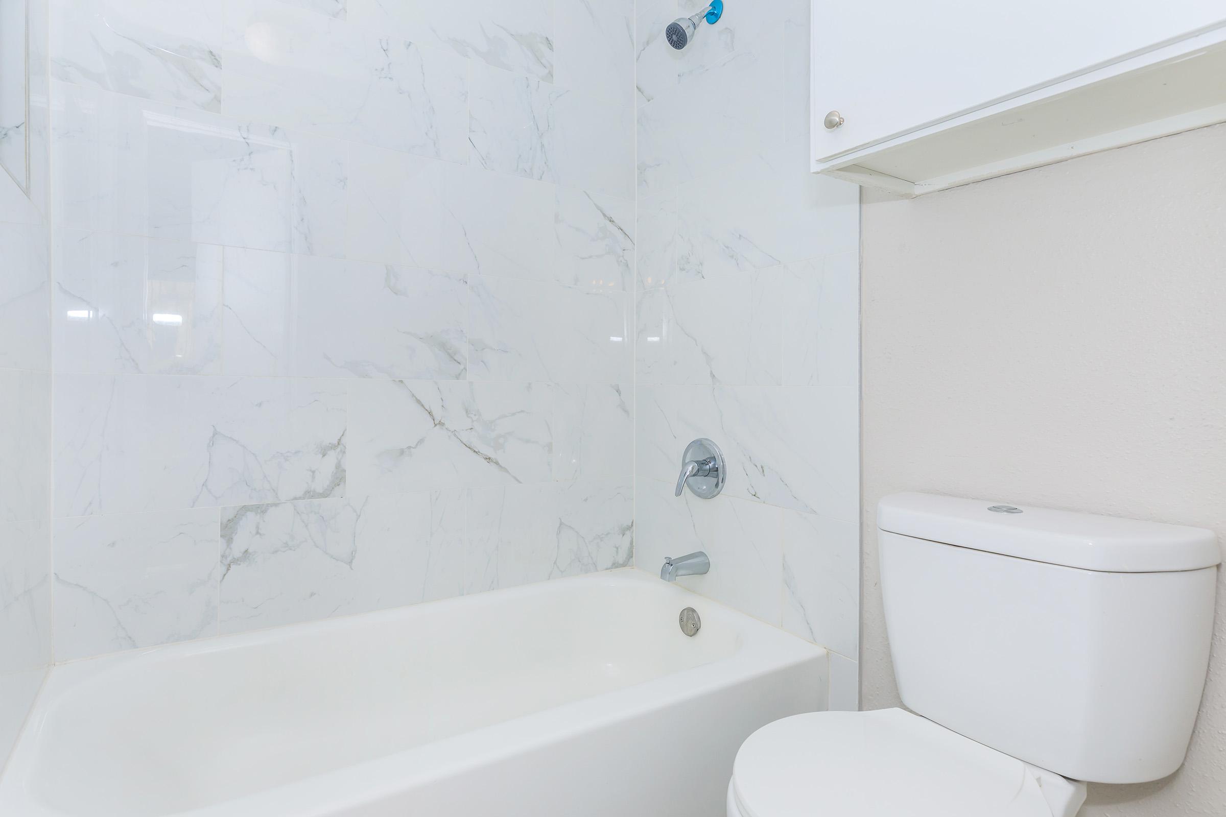 A clean and modern bathroom featuring a white bathtub with a marble-patterned wall behind it. There is a white toilet next to the bathtub, and a cabinet above with a simple, minimalist design. The overall aesthetic is light and spacious.