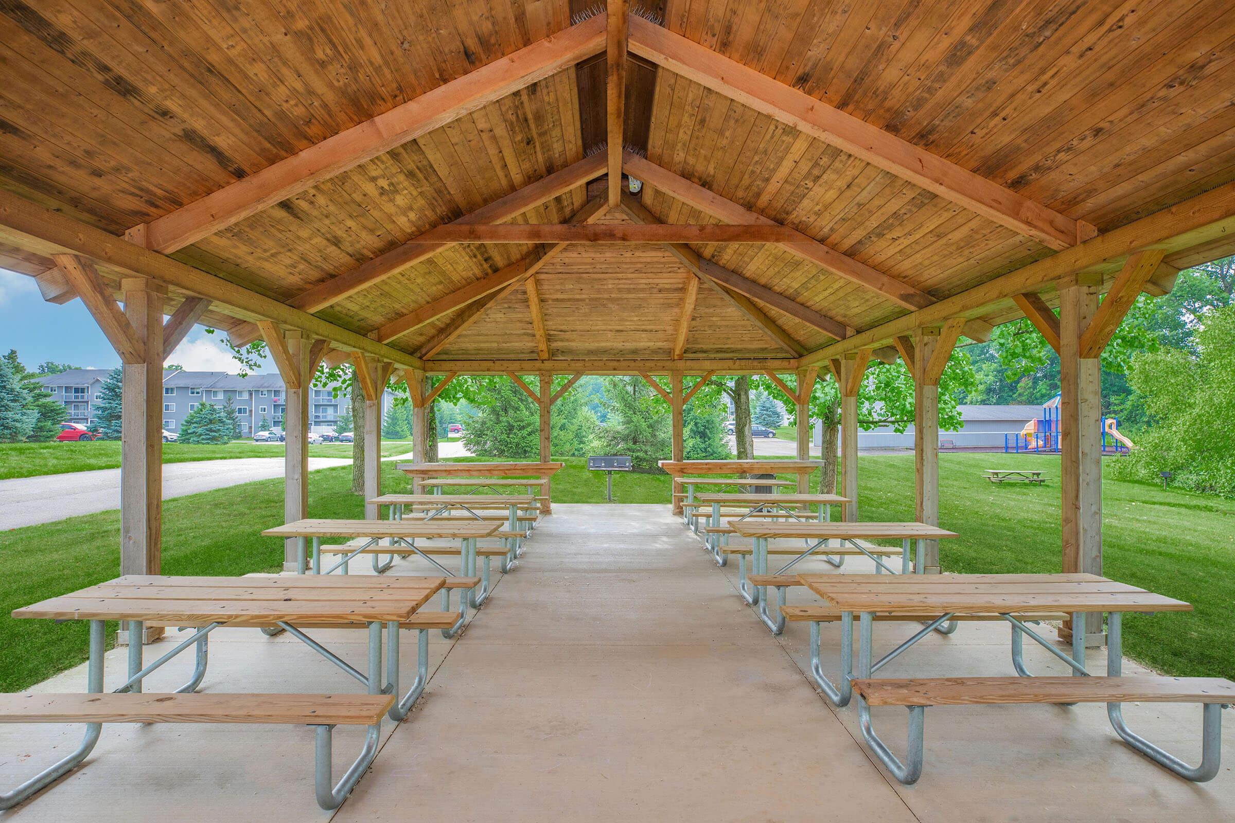 A spacious outdoor pavilion with wooden beams and a peaked roof. Several metal picnic tables are arranged neatly on a concrete floor, surrounded by lush green grass and trees. In the background, a pathway leads to a playground area. The atmosphere is bright and inviting, ideal for gatherings or events.