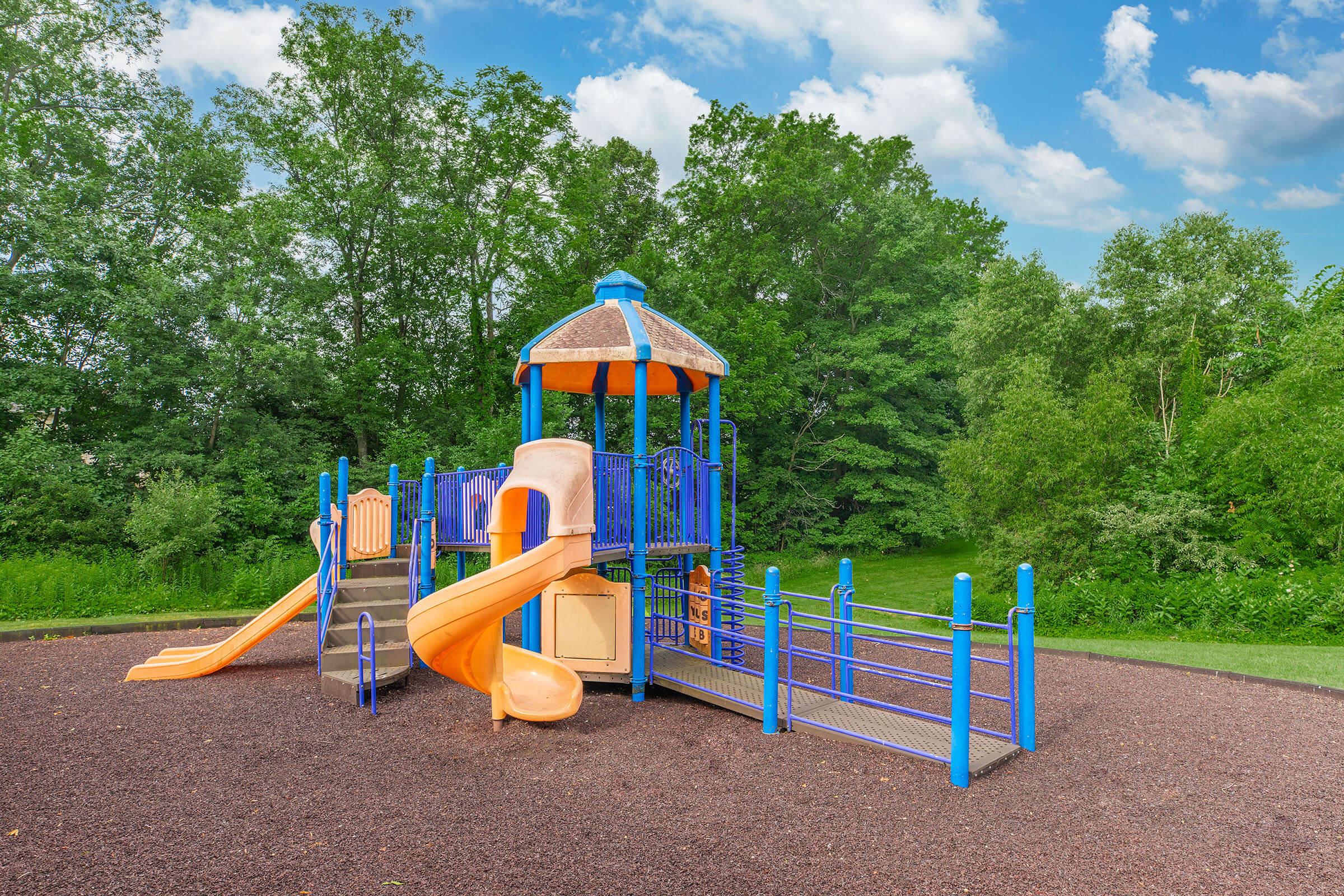A colorful playground structure featuring a slide, climbing areas, and a tower, surrounded by green trees against a bright blue sky with fluffy clouds. The play area is made of various vibrant colors like orange and blue, with soft ground cover for safety.