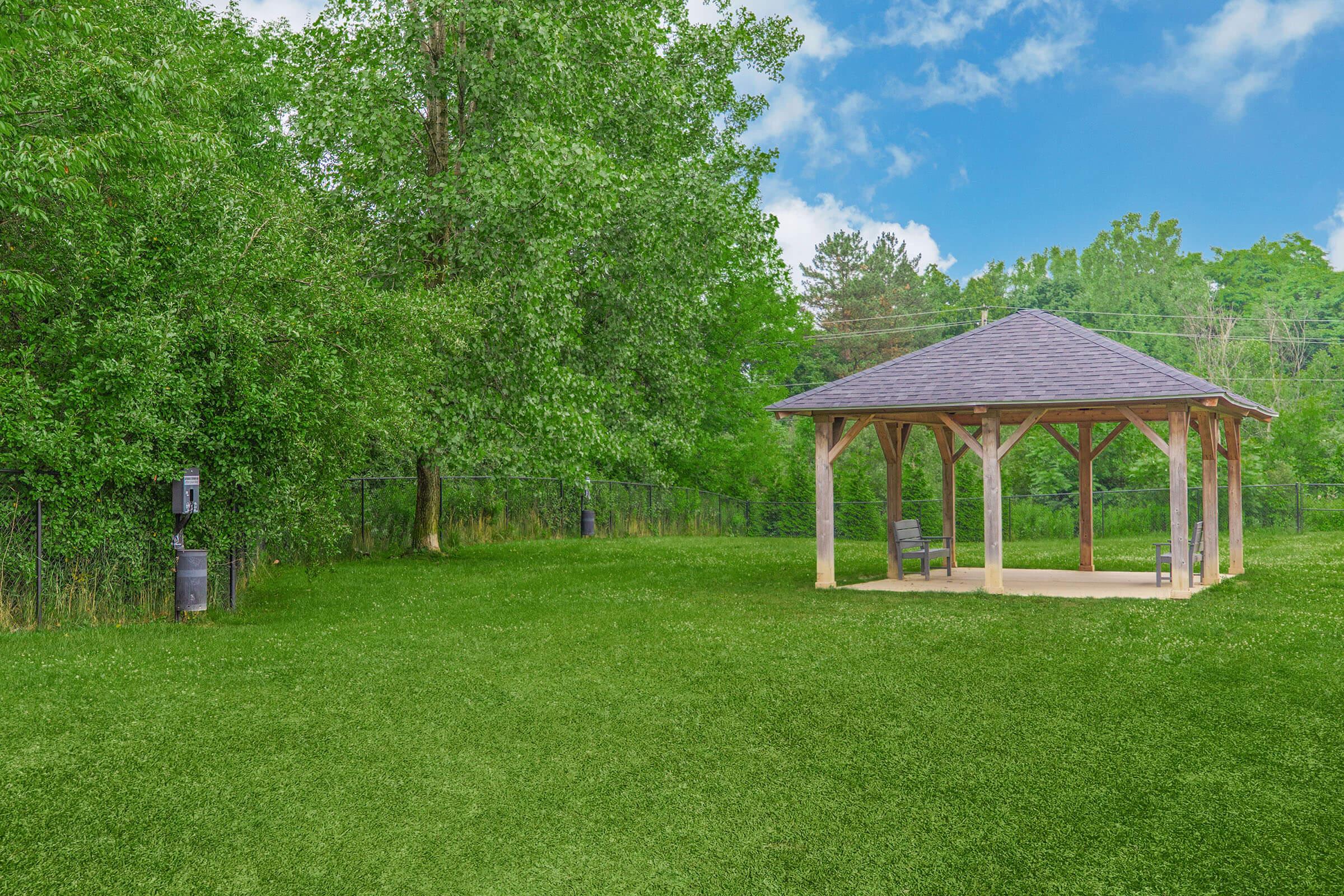 A spacious park scene featuring a wooden pavilion with a sloped roof, positioned on a lush green lawn. Surrounding the area are tall trees and a clear blue sky with scattered clouds, creating a tranquil and inviting outdoor environment.