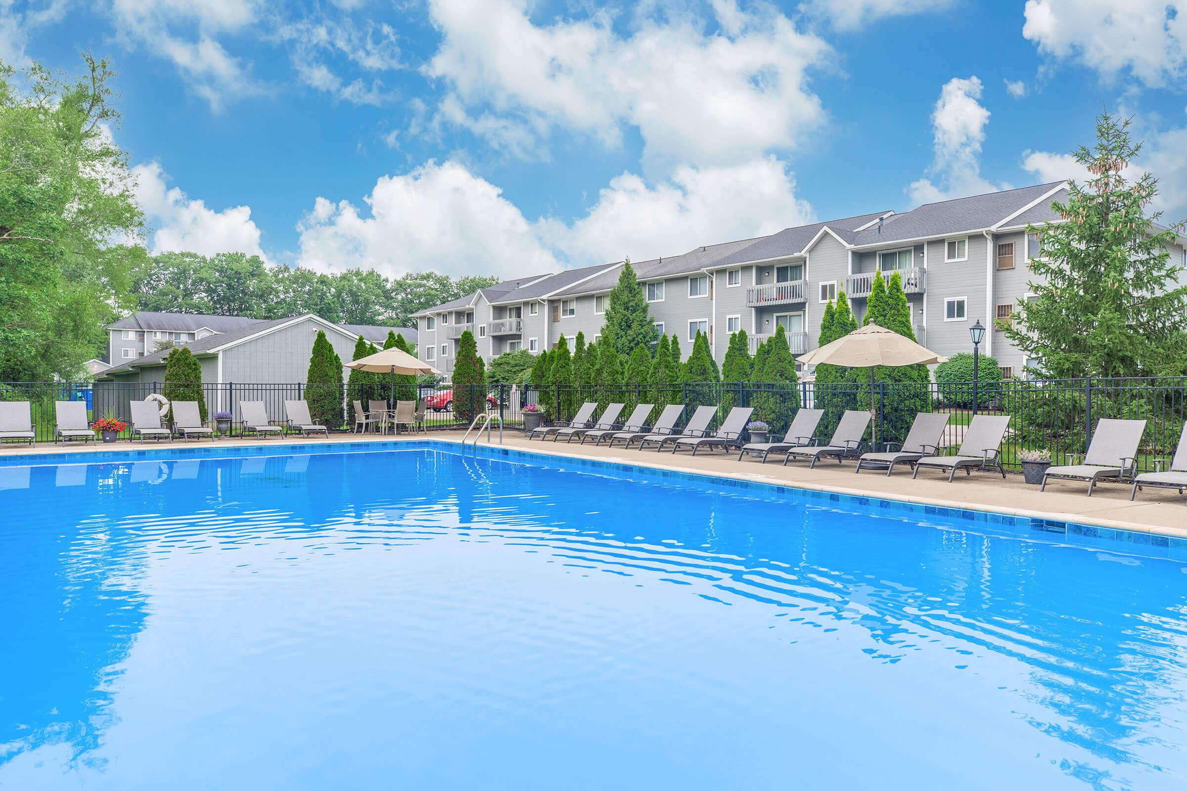 A large outdoor swimming pool surrounded by lounge chairs and umbrellas, with well-maintained greenery in the background. Apartment buildings are visible behind the pool, under a bright blue sky with fluffy white clouds.