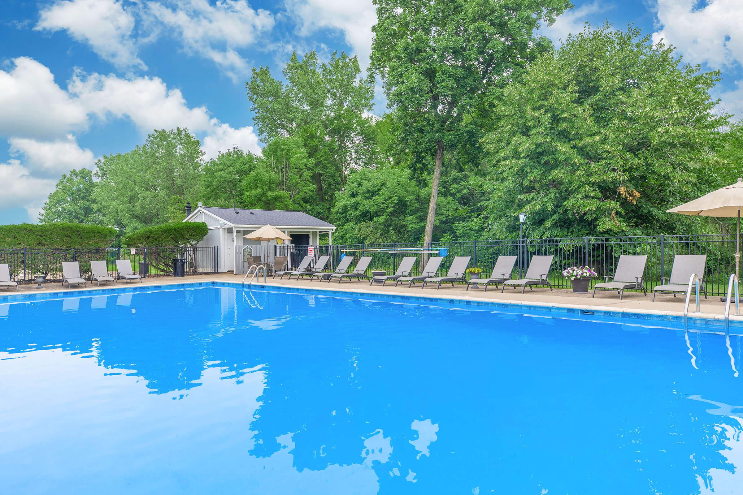 Swimming pool surrounded by lounge chairs and shaded umbrellas, with a backdrop of lush green trees and a cloudy blue sky. A small building is visible in the background, creating a serene and inviting atmosphere.