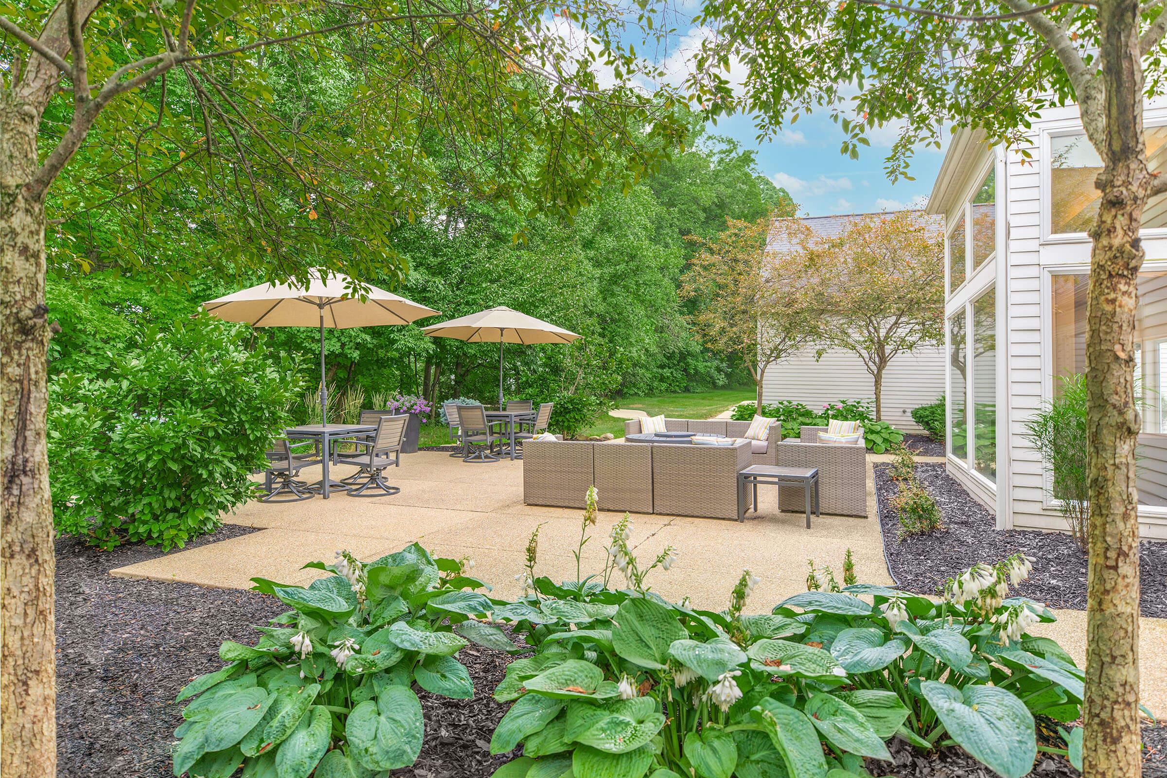 A serene outdoor patio area featuring light-colored furniture, large umbrellas, and surrounding greenery, including hostas and bushes. The space is designed for relaxation and entertainment, with a backdrop of trees and a modern home, creating a tranquil atmosphere.