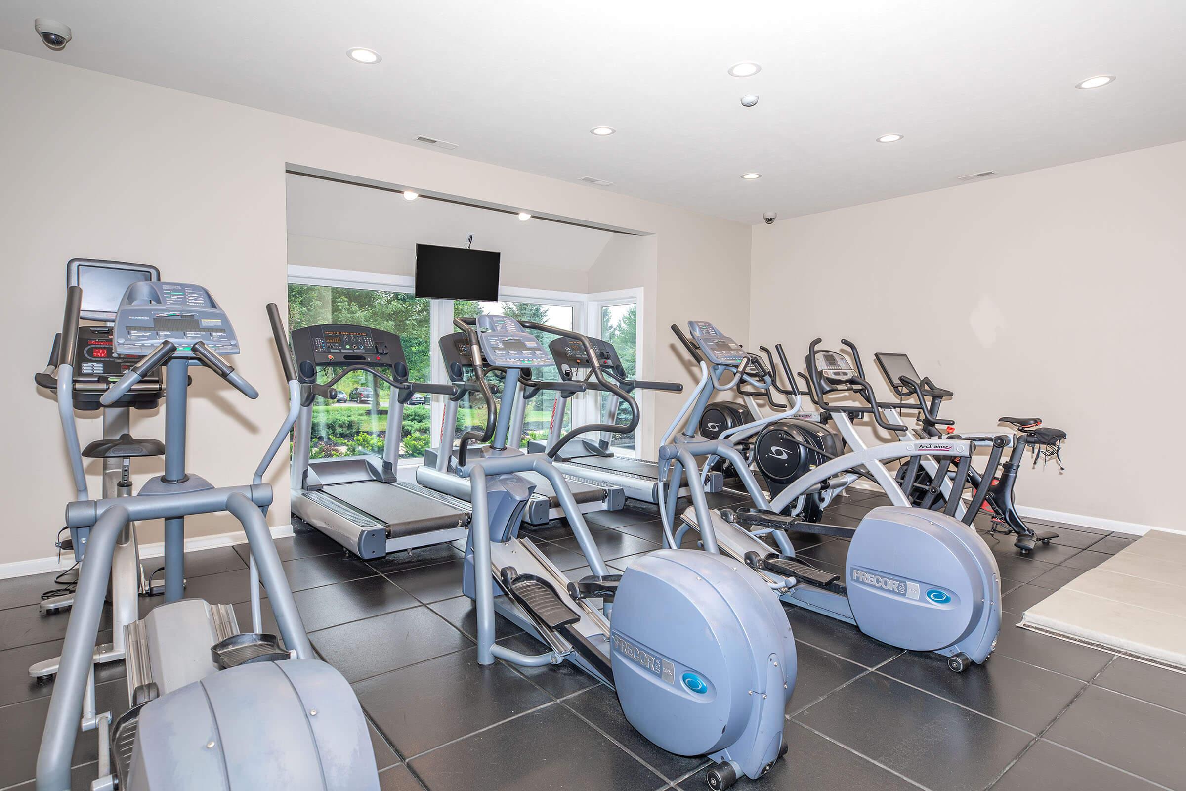 A modern fitness room featuring several pieces of exercise equipment, including treadmills, ellipticals, and stationary bikes. Large windows let in natural light, and a flat-screen TV is mounted on the wall. The floor is tiled, creating a clean and spacious workout environment.