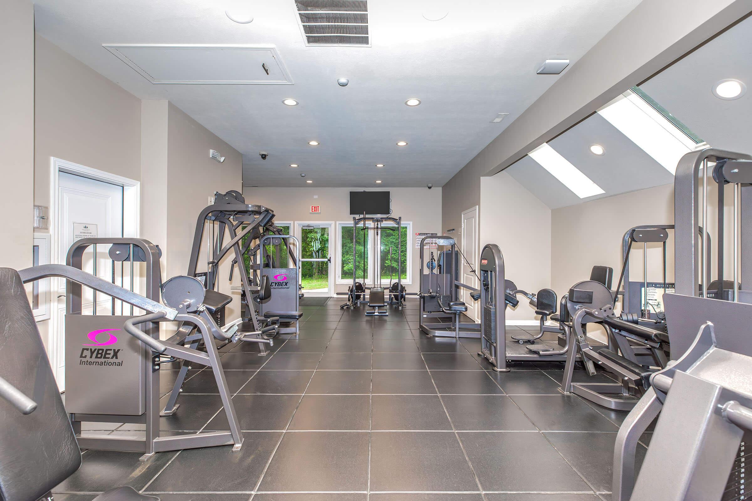 A modern gym interior featuring various exercise machines, including weight training equipment from Cybex. The space is well-lit with large windows, showcasing a clean and spacious layout, designed for fitness enthusiasts.