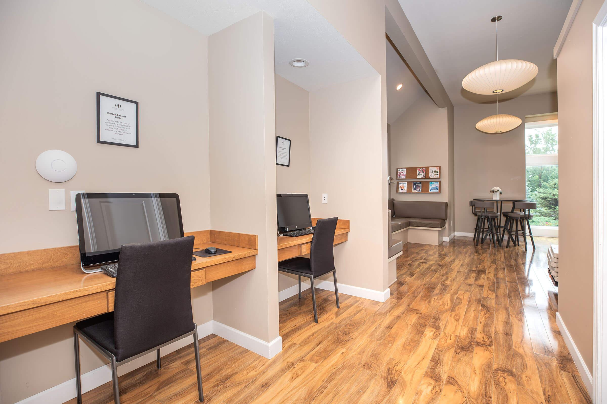 Two home office desks with computers, set against a light wall. Each desk has a black chair. The room features wooden floors and soft lighting. In the background, there is a seating area with a dining table and chairs, and large windows letting in natural light. Family photos adorn the walls.