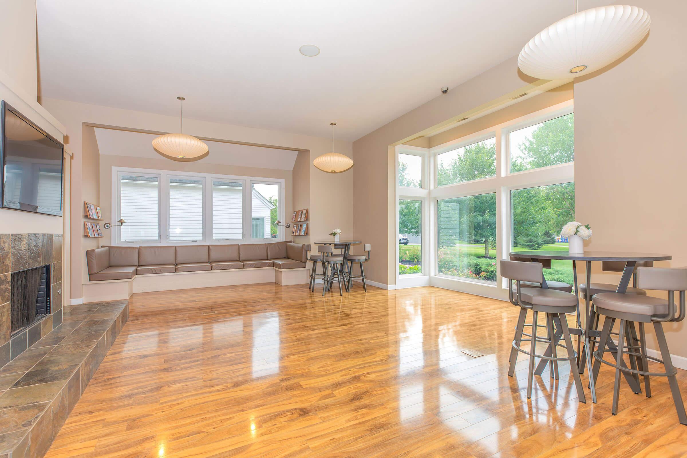 Modern interior of a spacious living area featuring hardwood flooring, large windows that let in natural light, and contemporary furniture. There are two stylish pendant lights, a comfortable seating arrangement along the wall, and a small dining table with chairs, creating an inviting atmosphere.