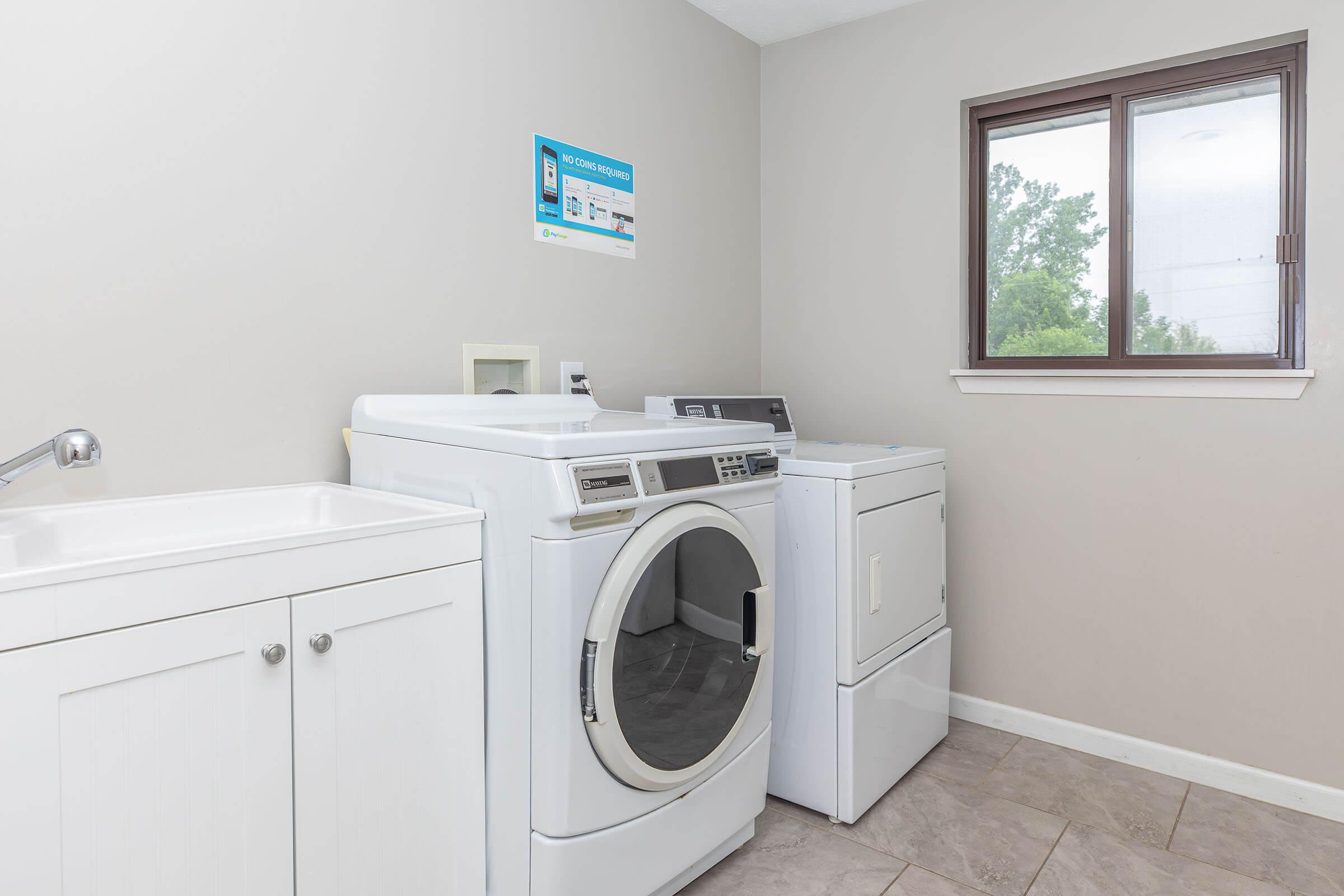 A bright laundry room featuring a white washer and dryer, a utility sink with cabinet storage, and a window letting in natural light. The walls are painted a light shade, and there’s a wall-mounted sign visible above the appliances, providing laundry instructions or information.