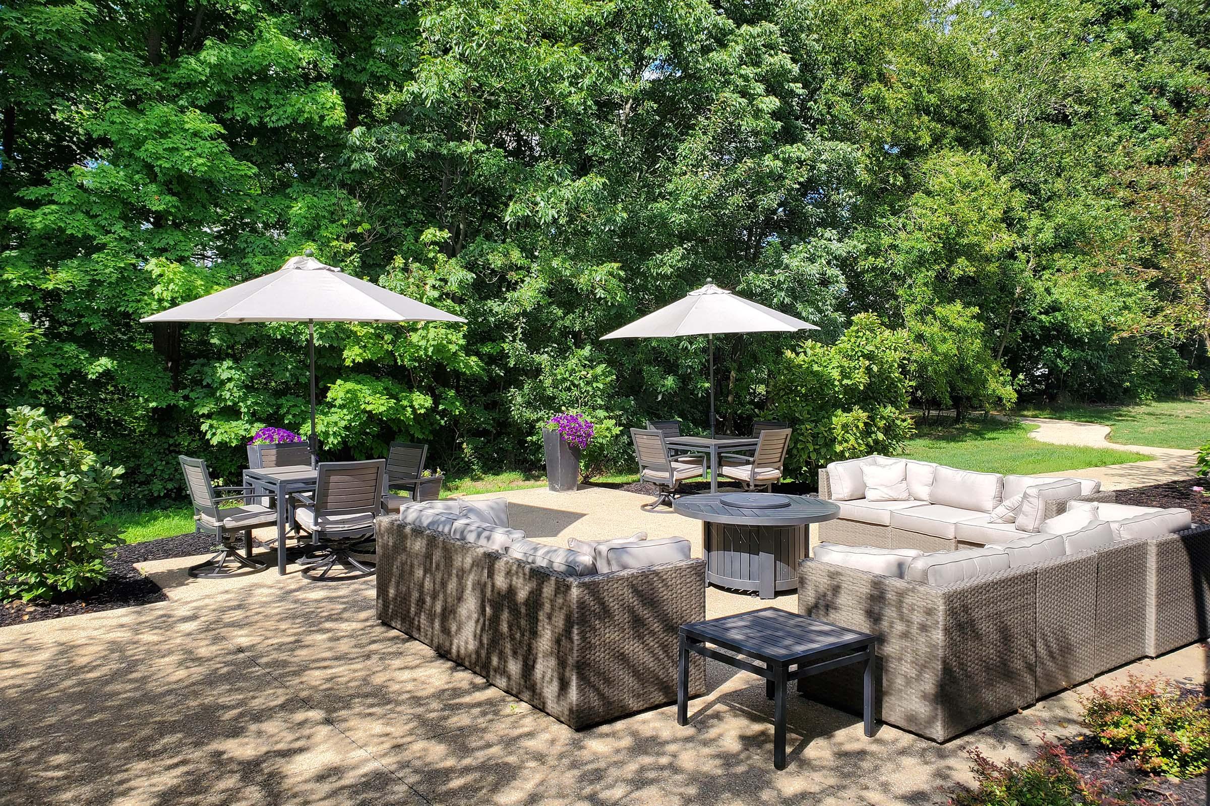 A cozy outdoor patio area featuring comfortable sectional seating and a coffee table, surrounded by lush greenery. Several patio tables with umbrellas provide shade, along with blooming plants adding a touch of color. The setting is inviting and ideal for relaxation or social gatherings.