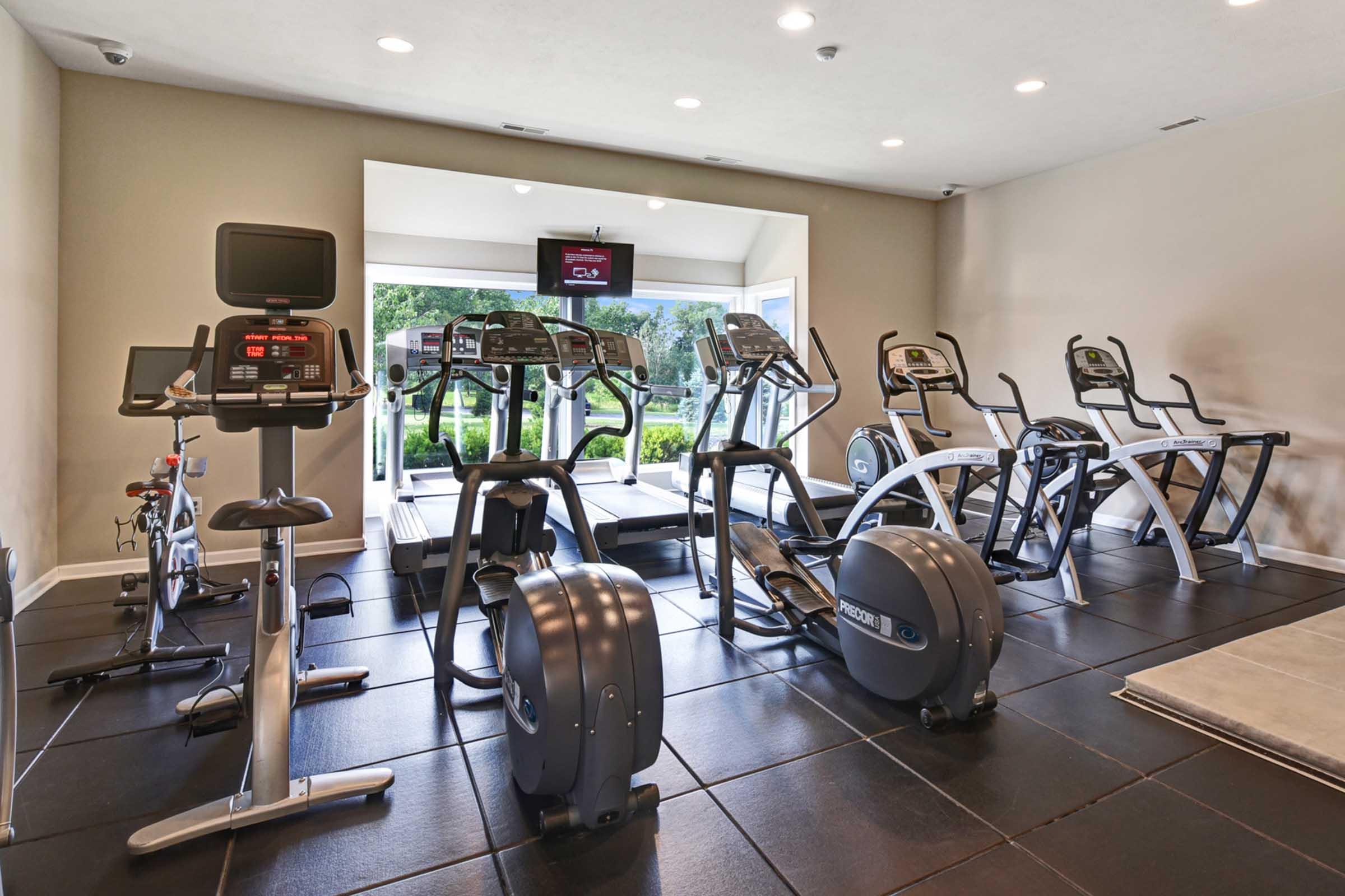 A modern gym featuring several cardio machines, including treadmills and stationary bikes. Large windows provide natural light and a view of greenery outside. The interior has a sleek, minimalistic design with rubber flooring and various exercise equipment arranged for convenience.