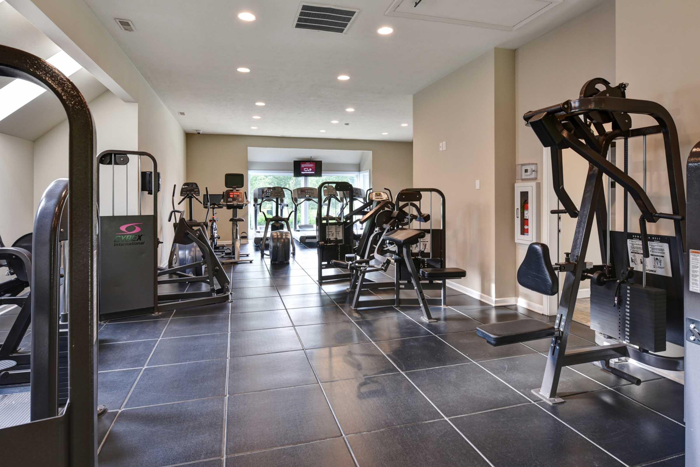 A modern gym interior featuring various workout machines, including weight training equipment and cardio machines, arranged in a spacious area with large windows allowing natural light. The floor is tiled, and the atmosphere is clean and organized, promoting a fitness-friendly environment.