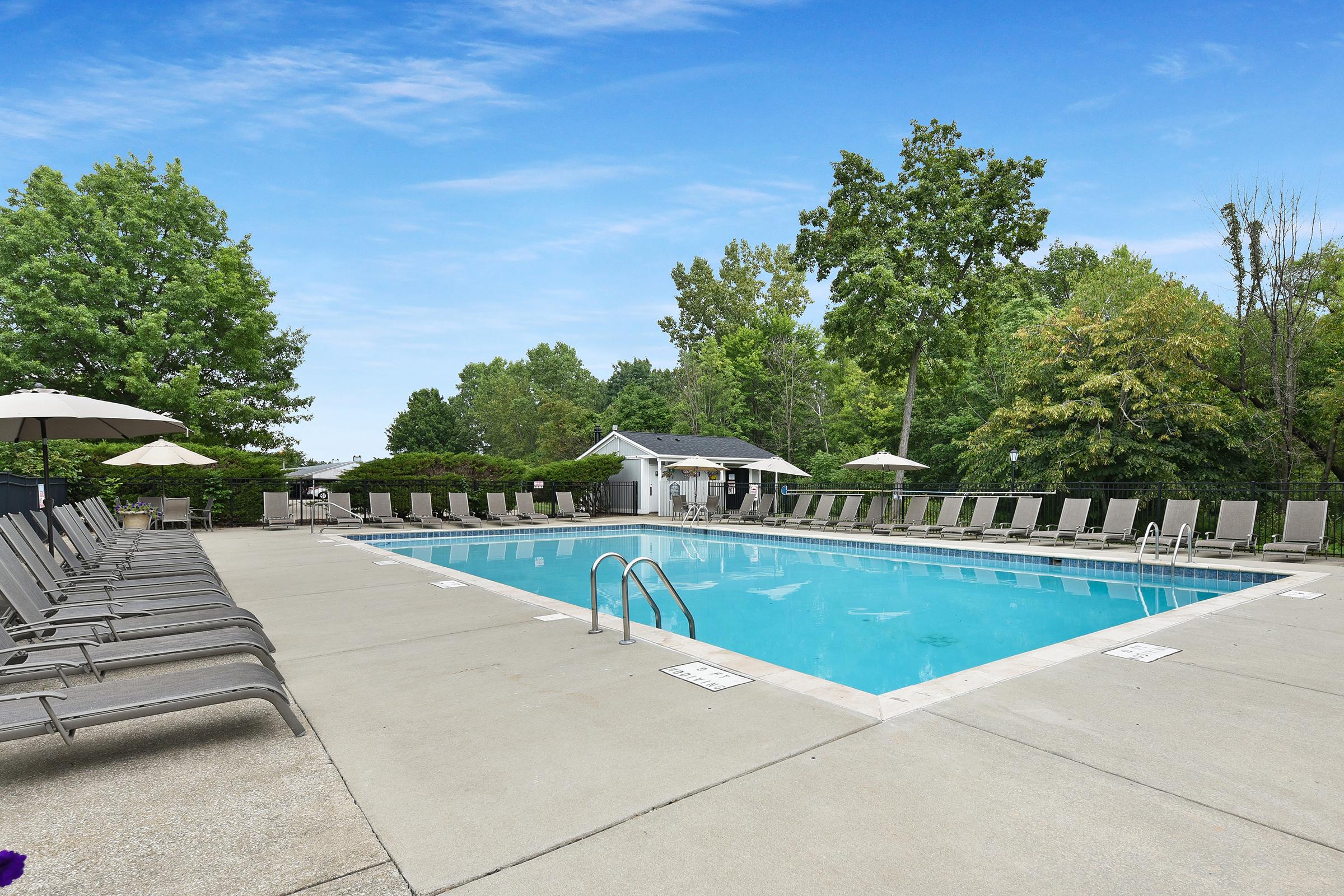 A clear outdoor swimming pool surrounded by lounge chairs, with a small white building and trees in the background. Umbrellas provide shade, and the pool area is well-maintained under a bright blue sky.