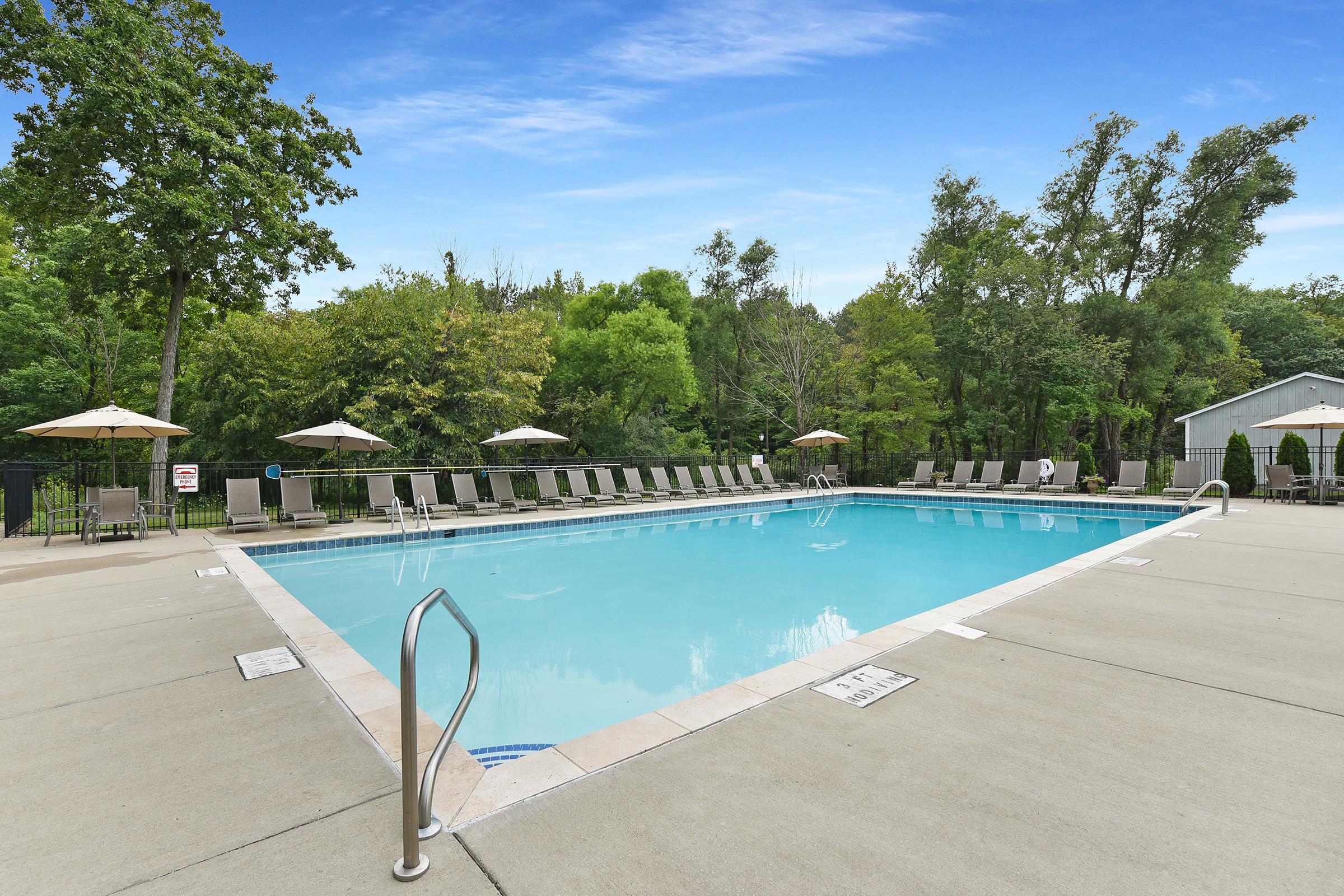 A clear swimming pool surrounded by lounge chairs and umbrellas, set in a tranquil outdoor environment with lush greenery and trees in the background. The sky is bright and blue, reflecting a peaceful and inviting atmosphere.
