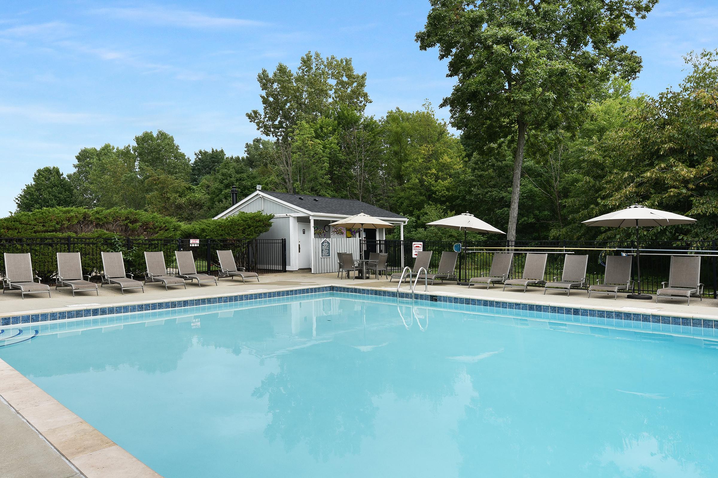 A serene swimming pool surrounded by lounge chairs and umbrellas, with lush greenery and a small building in the background. The water is crystal clear under a bright blue sky, creating a relaxing outdoor atmosphere perfect for leisure and enjoyment.