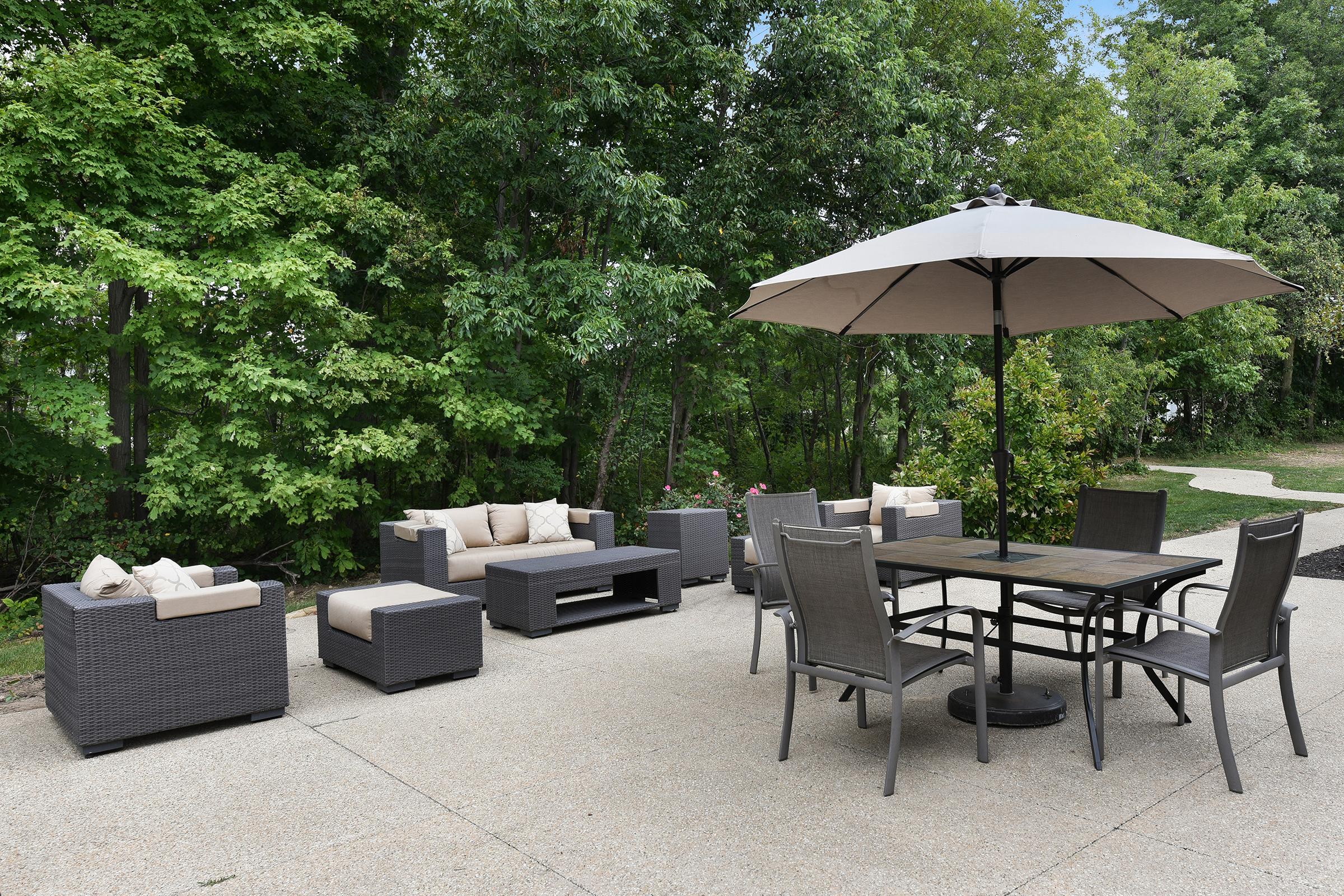 Outdoor patio area featuring a set of gray wicker furniture with beige cushions, including a seating arrangement with two armchairs and a sofa, a coffee table, and a dining table with metal chairs. A large umbrella provides shade, surrounded by lush green trees.