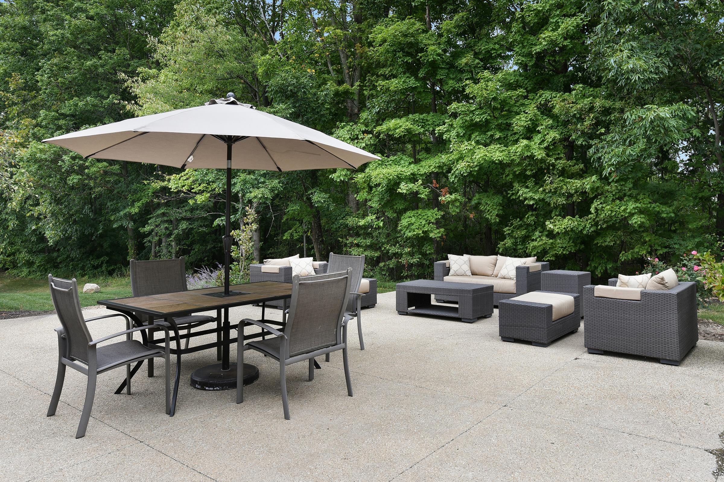 A spacious outdoor patio featuring a wooden dining table with six metal chairs under a large umbrella, and a cozy seating area with cushioned wicker sofas and a coffee table, surrounded by lush greenery.
