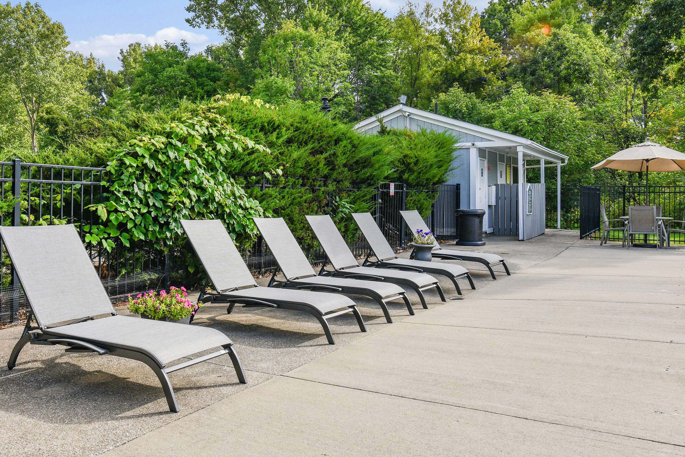 A row of gray lounge chairs lined up by a pool area, surrounded by greenery and flowering plants. In the background, a small building is visible, along with an umbrella and additional seating, creating a relaxing outdoor space.