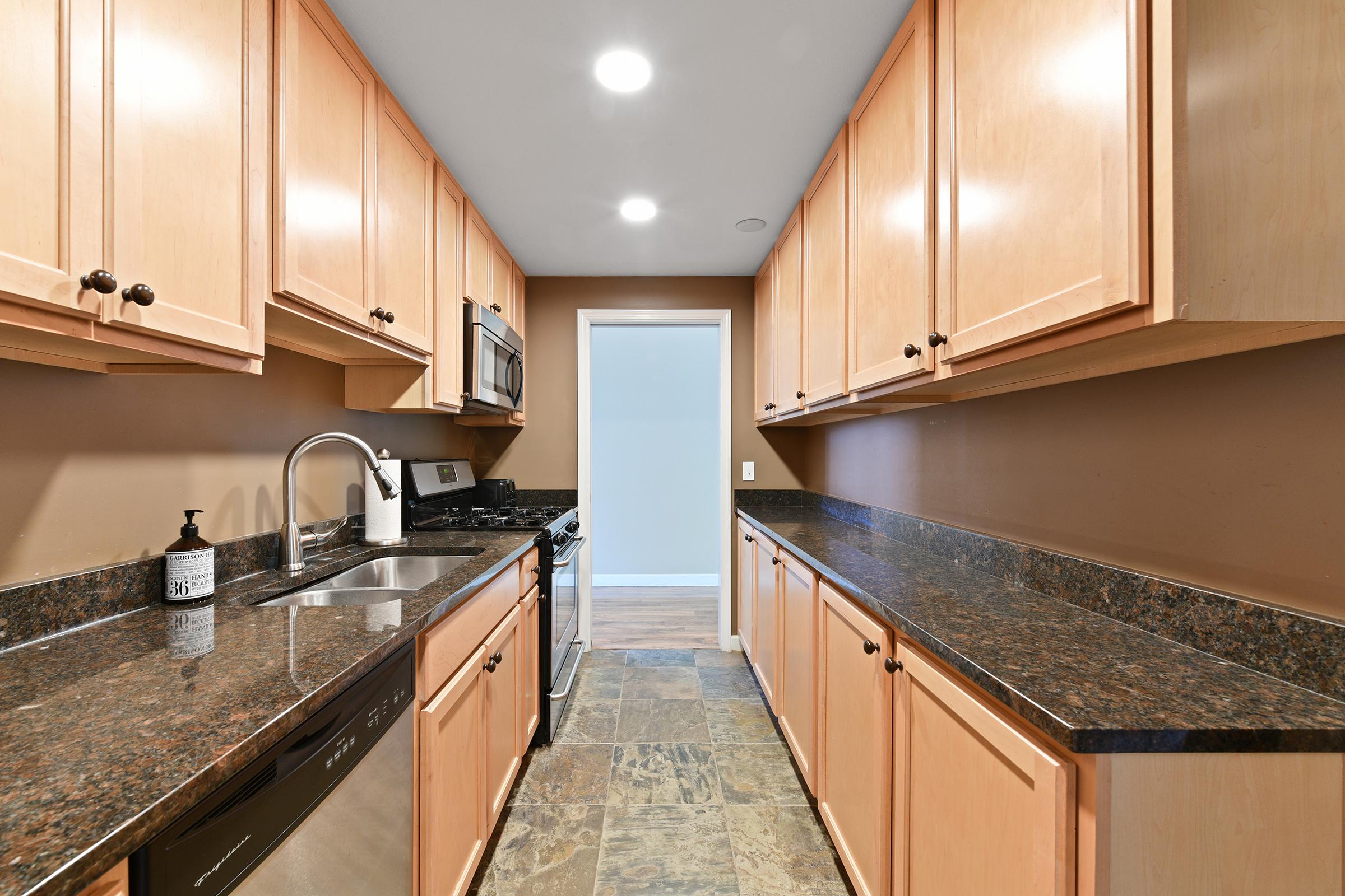 A modern kitchen featuring light wood cabinetry, dark granite countertops, stainless steel appliances including a microwave and dishwasher, and a sleek sink. The walls are painted a warm neutral color, and there is a bright, well-lit atmosphere with overhead lights. A door leads to another room.