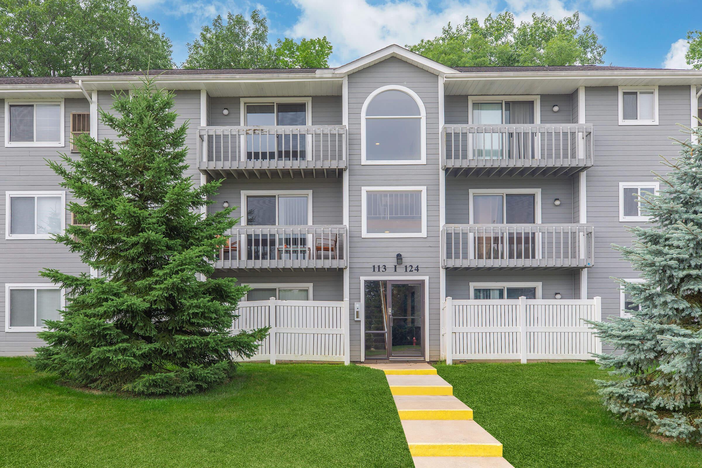 A modern, three-story apartment building with gray siding, featuring multiple balconies and large windows. In front, there is a neatly maintained lawn with two evergreen trees and a pathway leading to the entrance, framed by a white picket fence. The sky is partly cloudy.